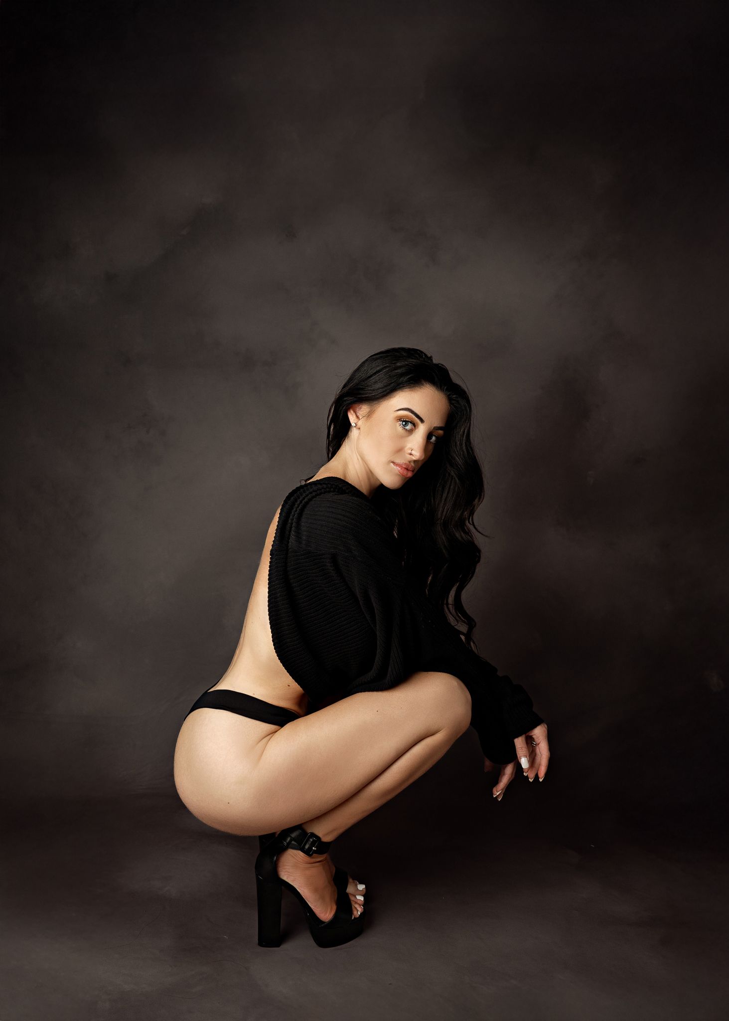Artistic fashion portrait of model in black sweater and heels crouching against dark studio backdrop.