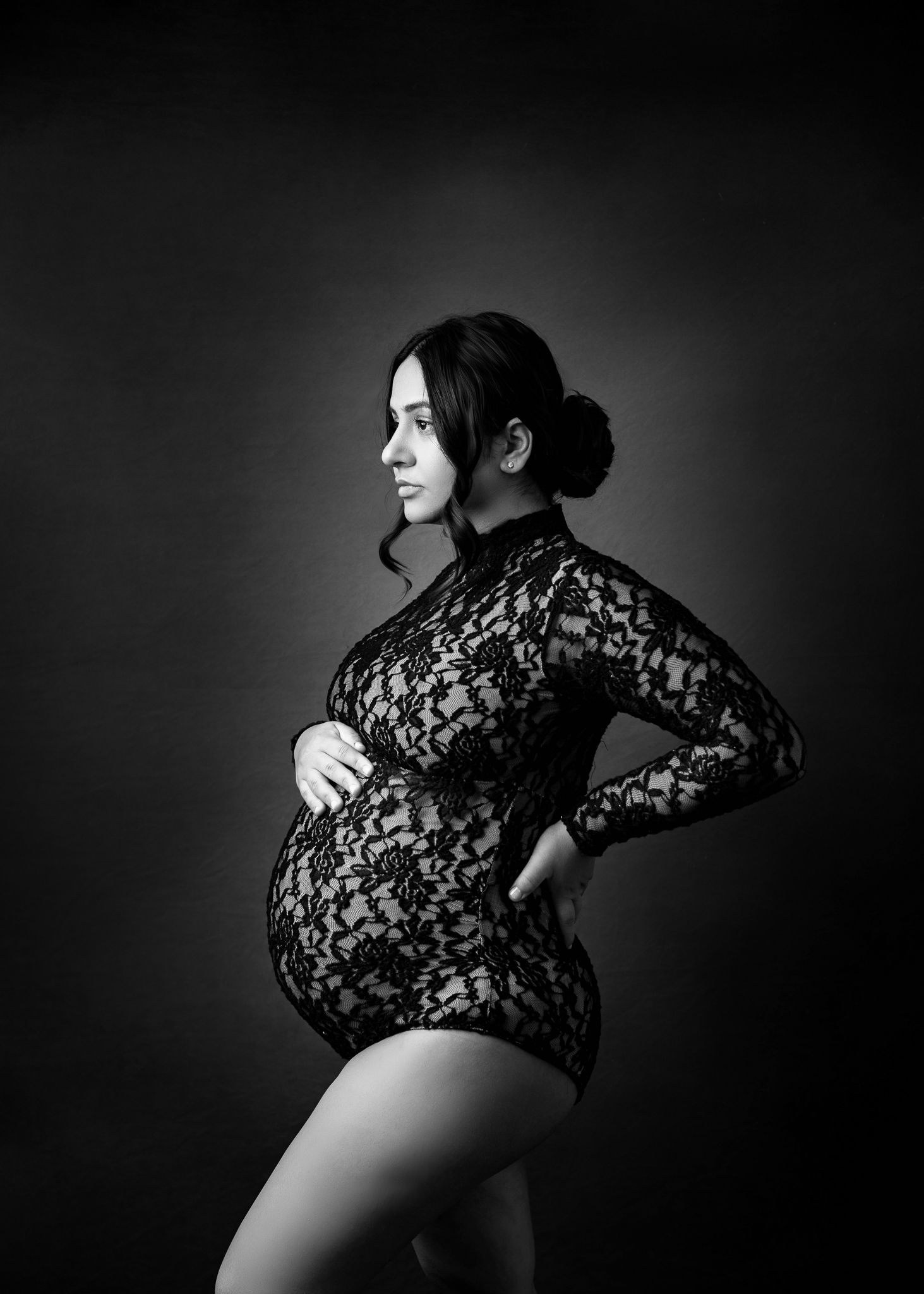 Black and white maternity photo showing a side profile pose in a lace bodysuit against a dark backdrop.