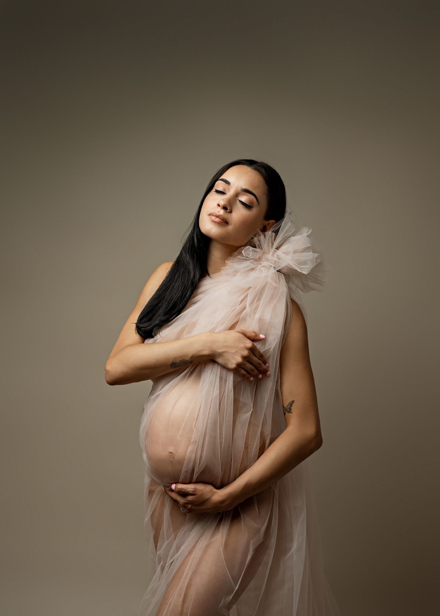 Woman in sheer beige tulle dress poses gracefully against neutral backdrop in artistic maternity photoshoot.