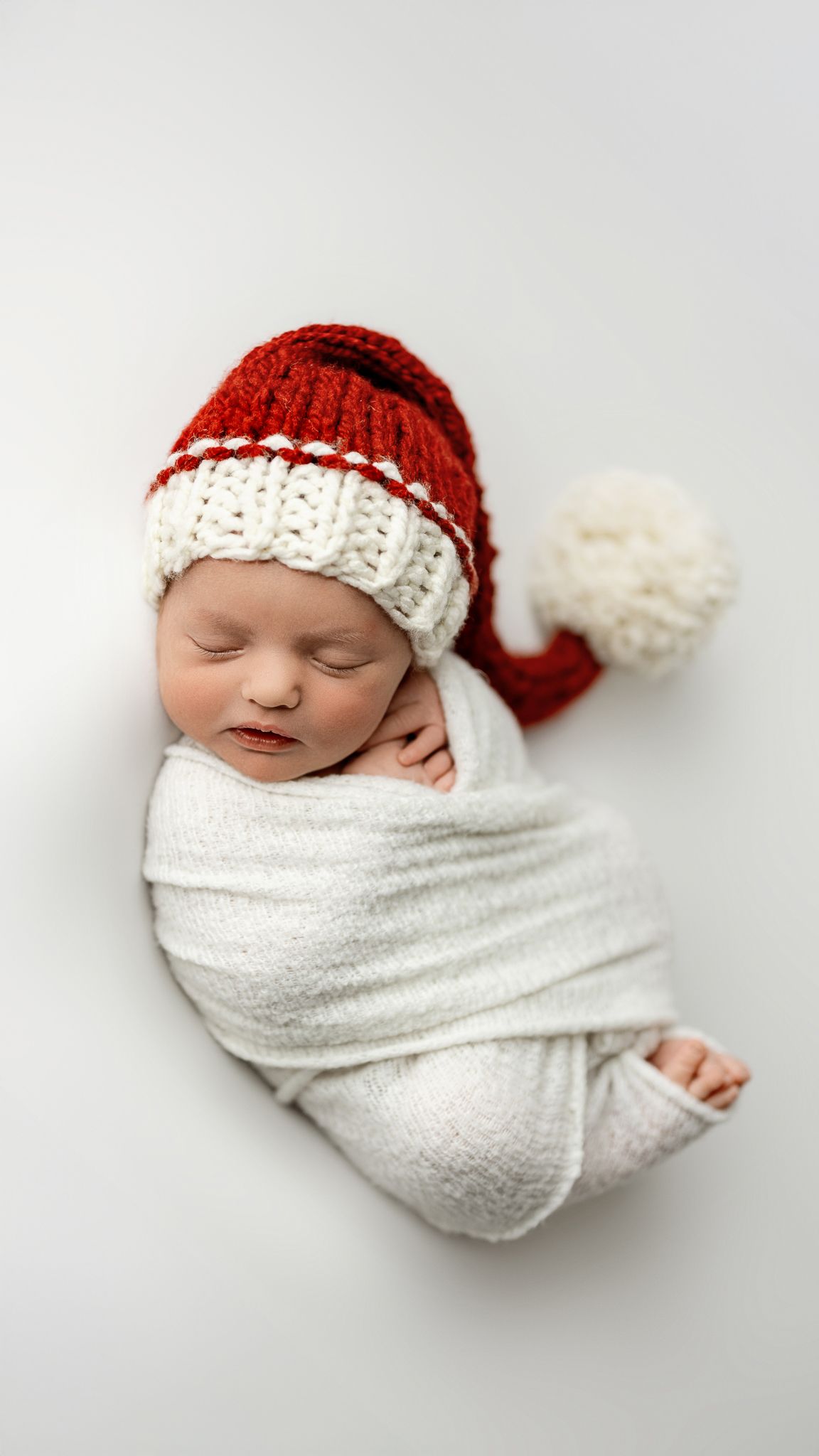 A sleeping newborn wrapped in white swaddle wearing a red and white knitted Santa hat against light background.