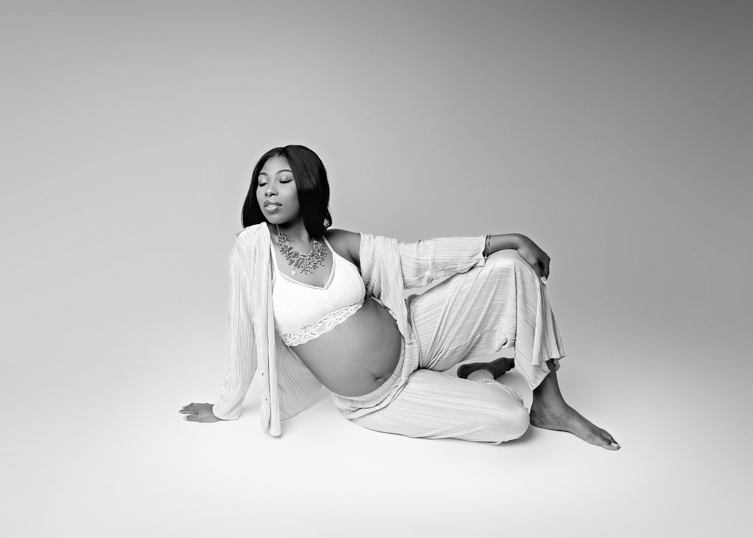 Black and white maternity portrait of someone wearing a white outfit posed on the floor in a studio setting.
