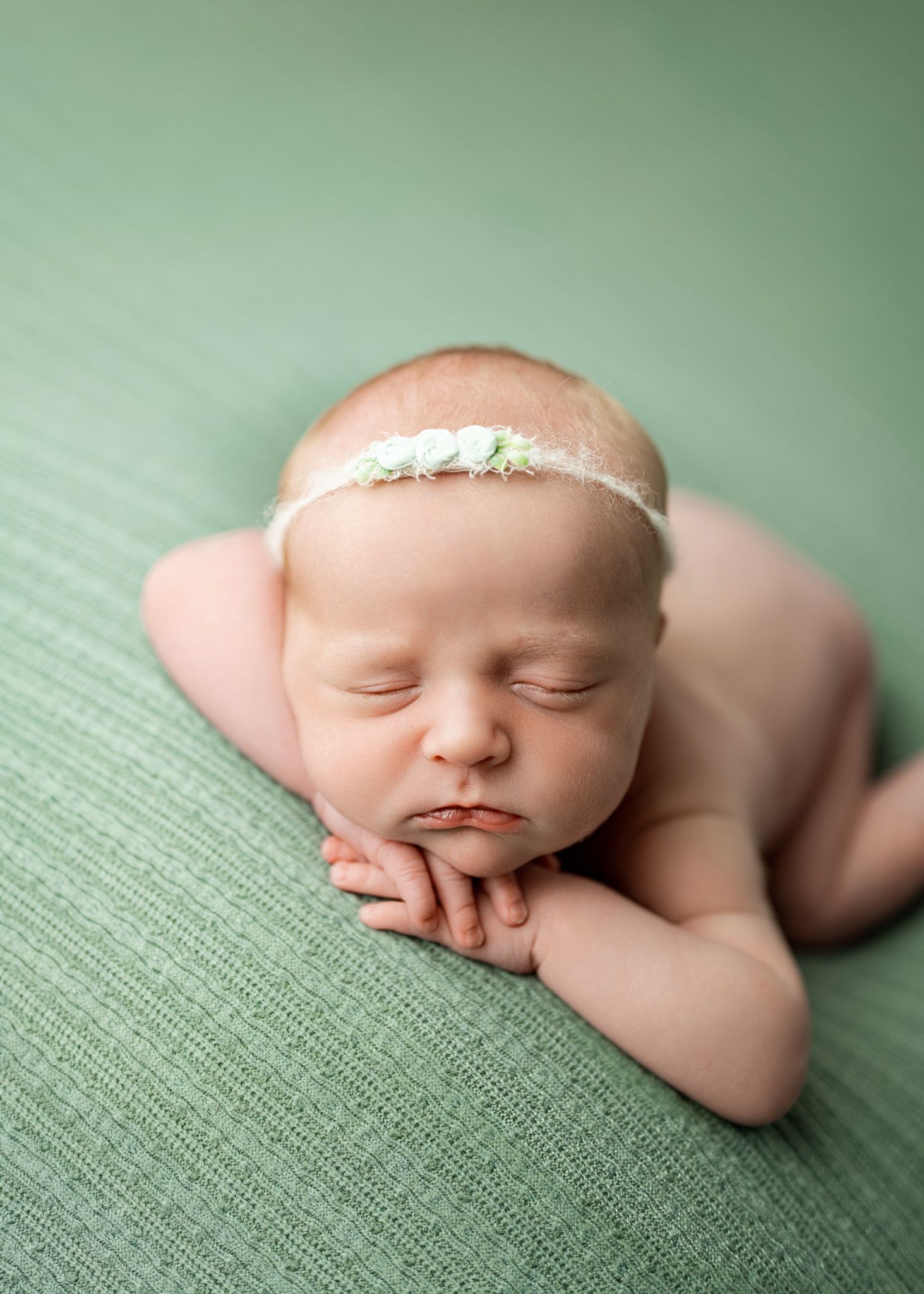A sleeping newborn swaddled on textured green fabric with a delicate lace headband accessory lies peacefully posed.