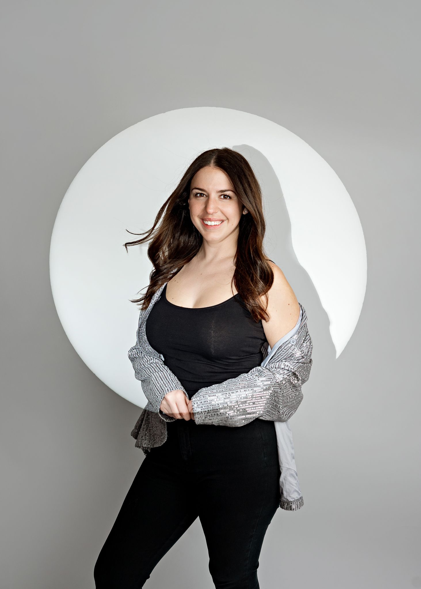 A woman in a black tank top and shiny silver cardigan poses confidently against a white circular backdrop on gray background.