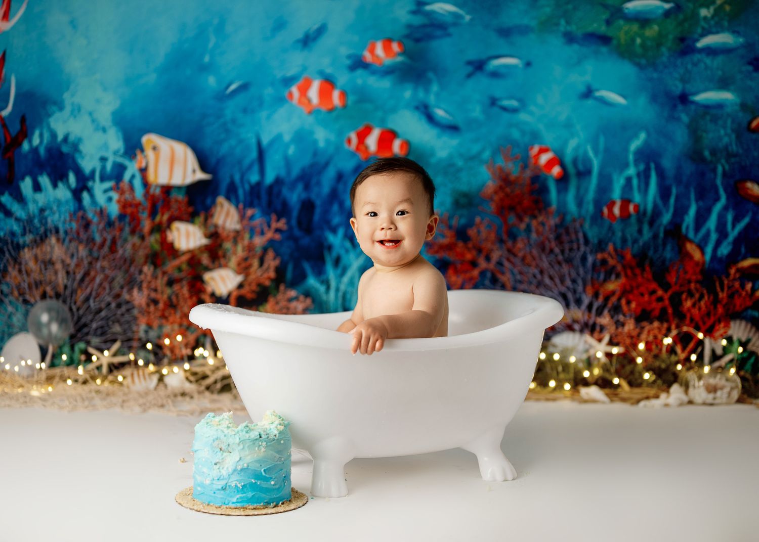 Baby sits in white bathtub prop with underwater ocean backdrop and blue smash cake for first birthday photo shoot.