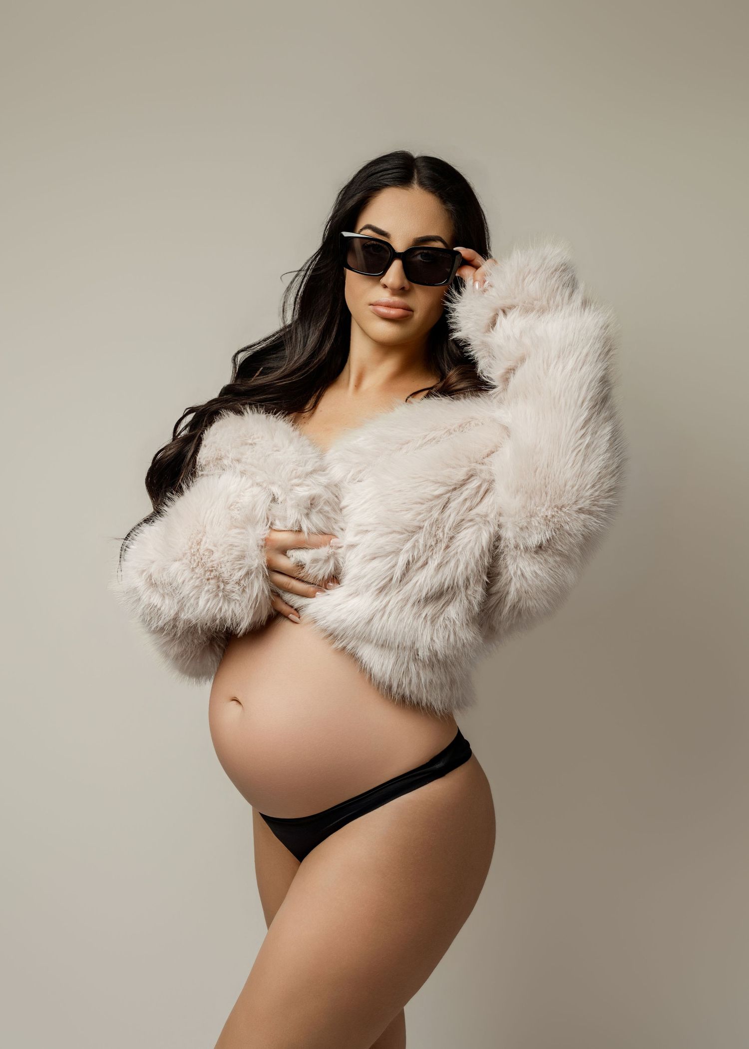 Pregnant woman in fluffy white faux fur jacket and black bikini bottoms wearing cat-eye sunglasses in a studio maternity photo.