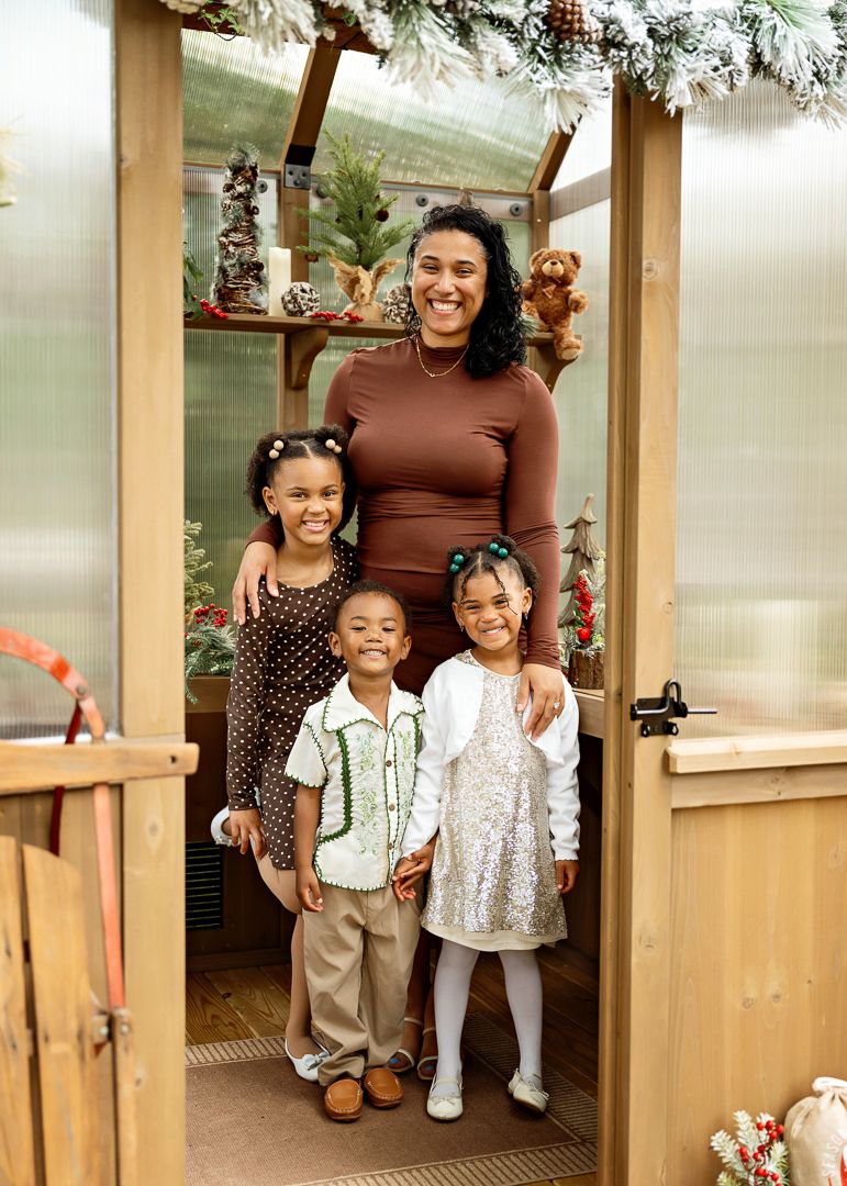 A smiling woman poses with three children in a festive greenhouse decorated with Christmas trees and holiday decor.