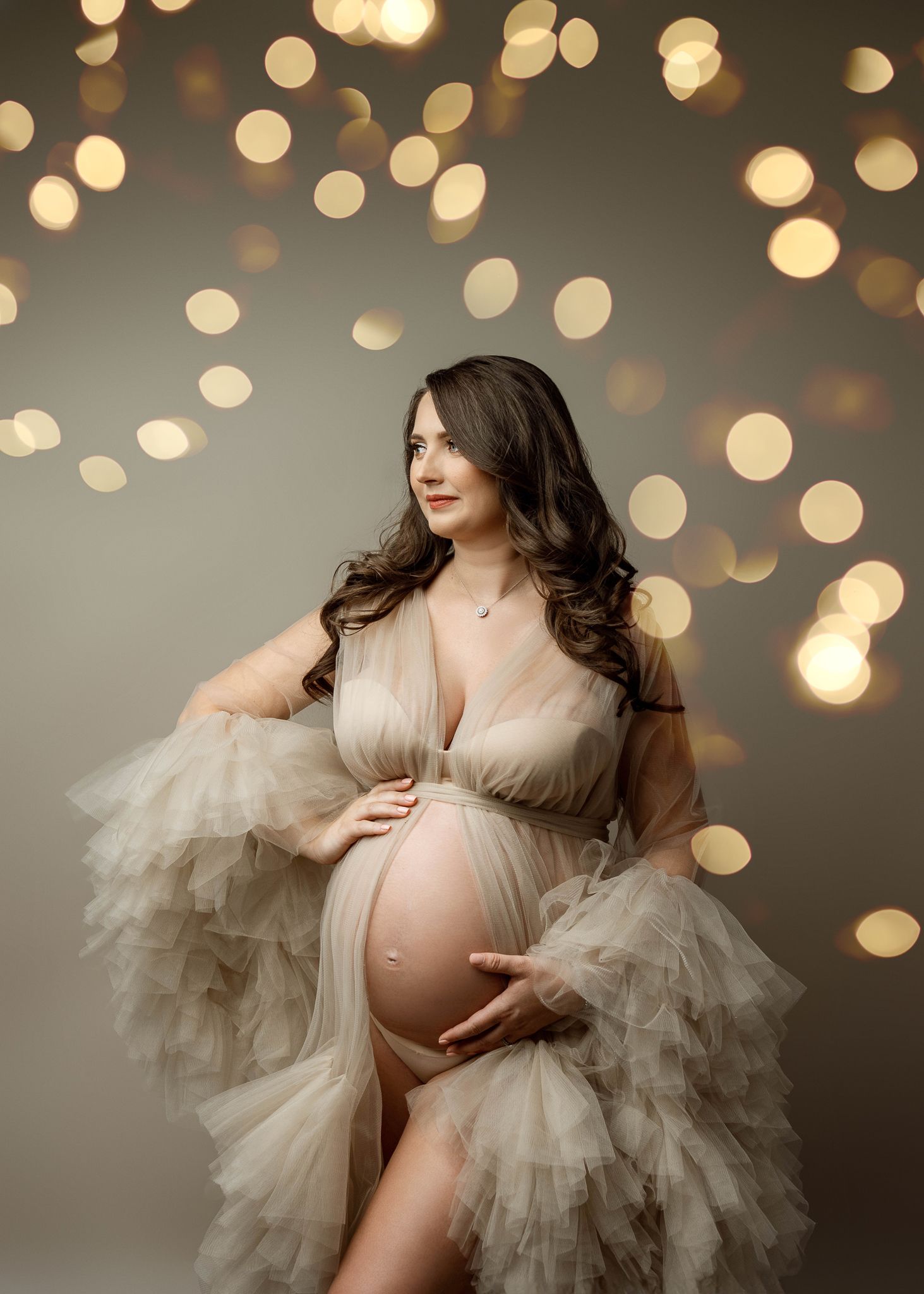 Elegant maternity photo with sheer tulle dress and romantic bokeh lights creating dreamy backdrop.