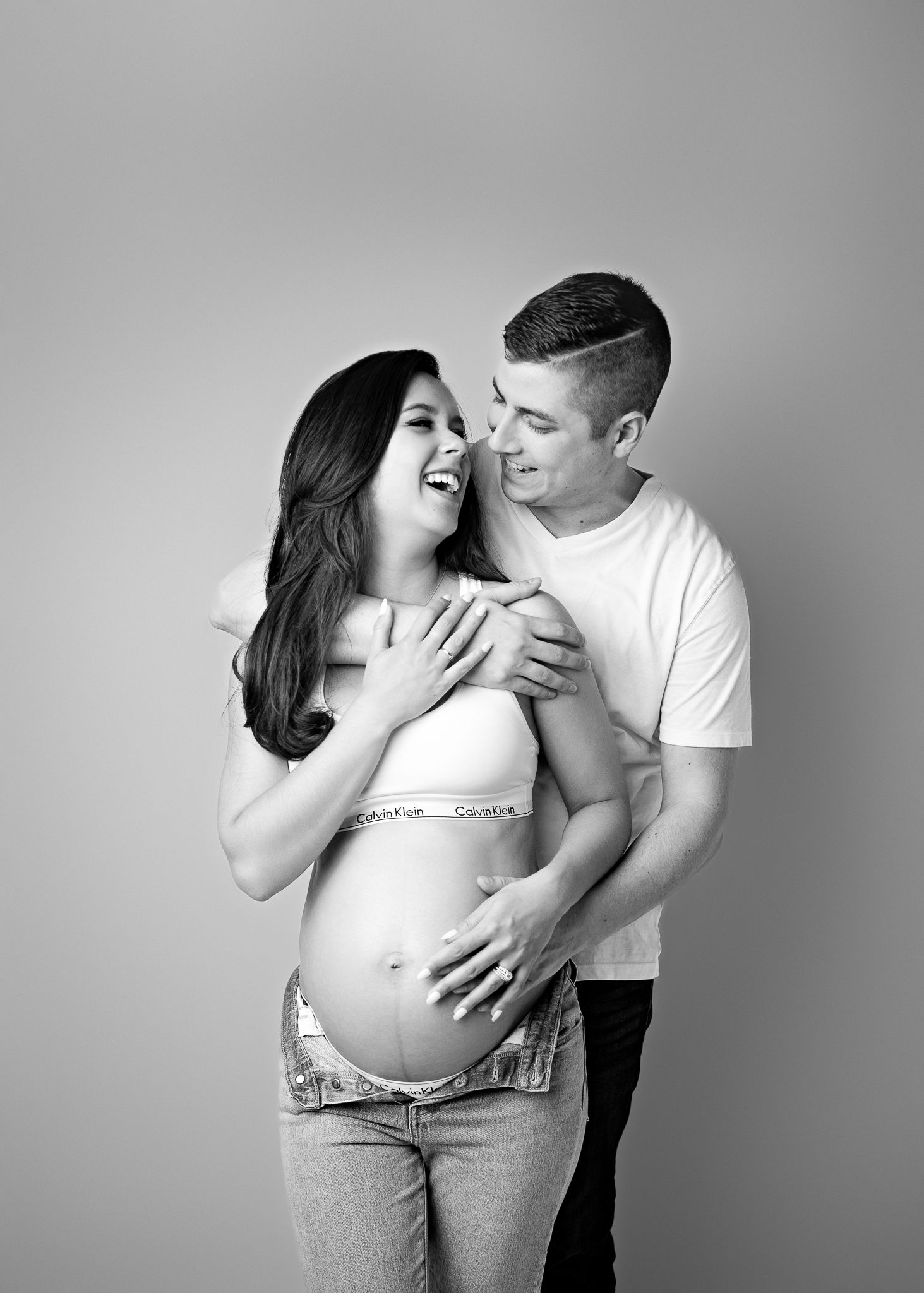 Black and white maternity photo of a couple in casual attire embracing and laughing against a plain background.