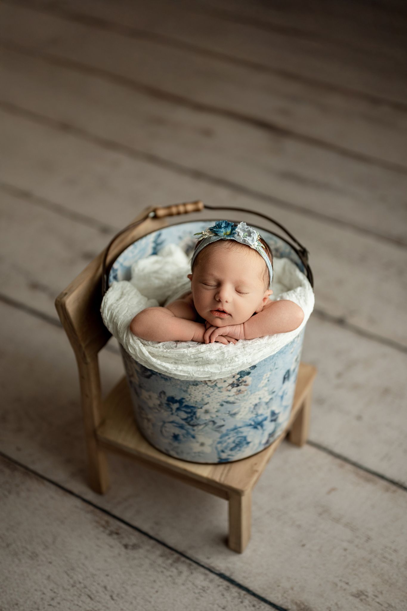 A sleeping newborn baby nestled in a blue and white floral basket on a rustic wooden floor.
