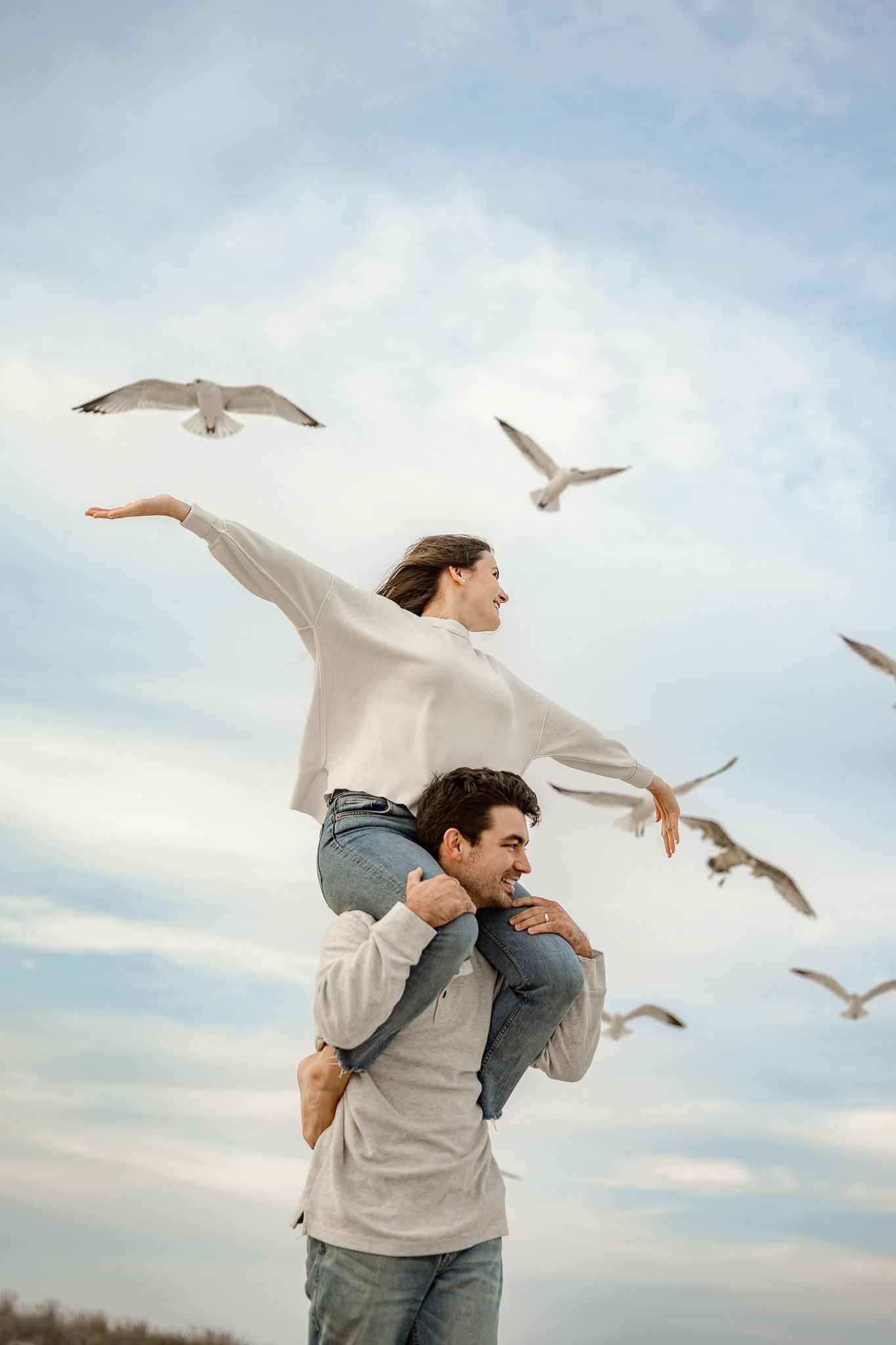 Two people in casual clothes dance and spin together outdoors with seagulls flying around them against a cloudy sky.