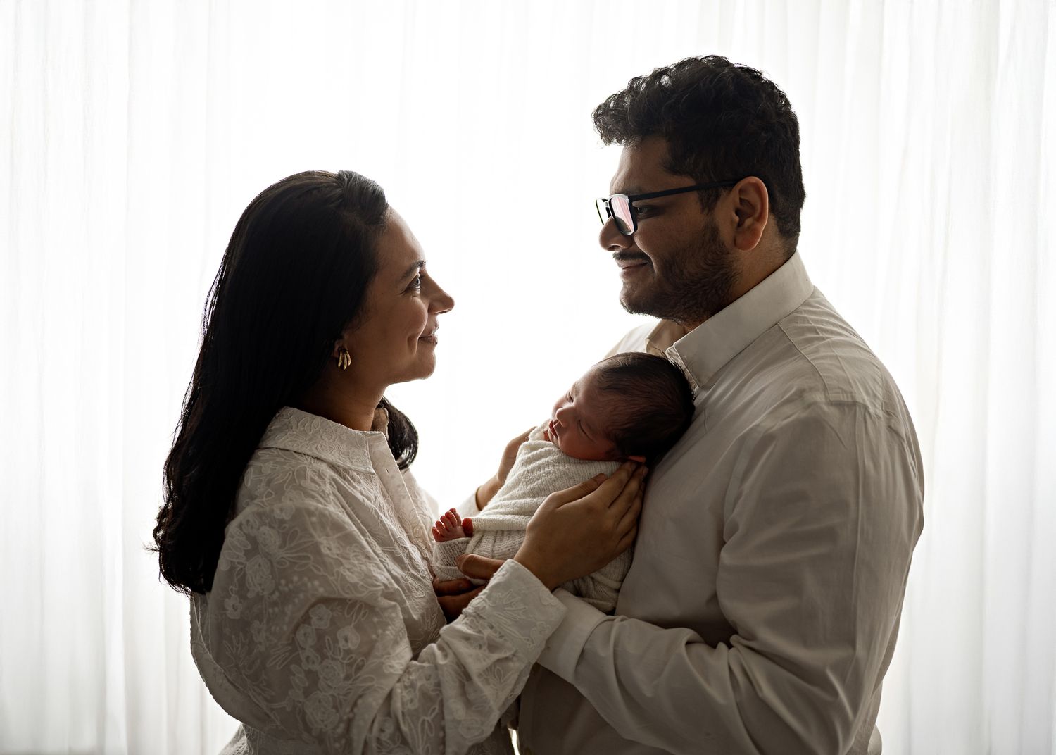 Silhouetted figures in white clothing share an intimate family moment while holding their baby against soft light from a window.