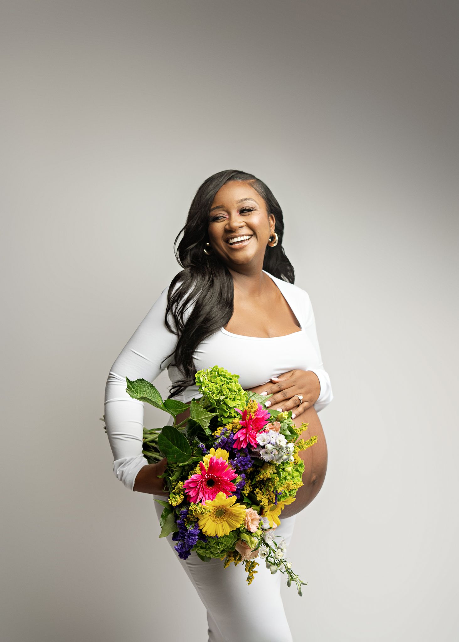 A pregnant person in a white dress holds a vibrant bouquet of colorful flowers during a maternity photoshoot.