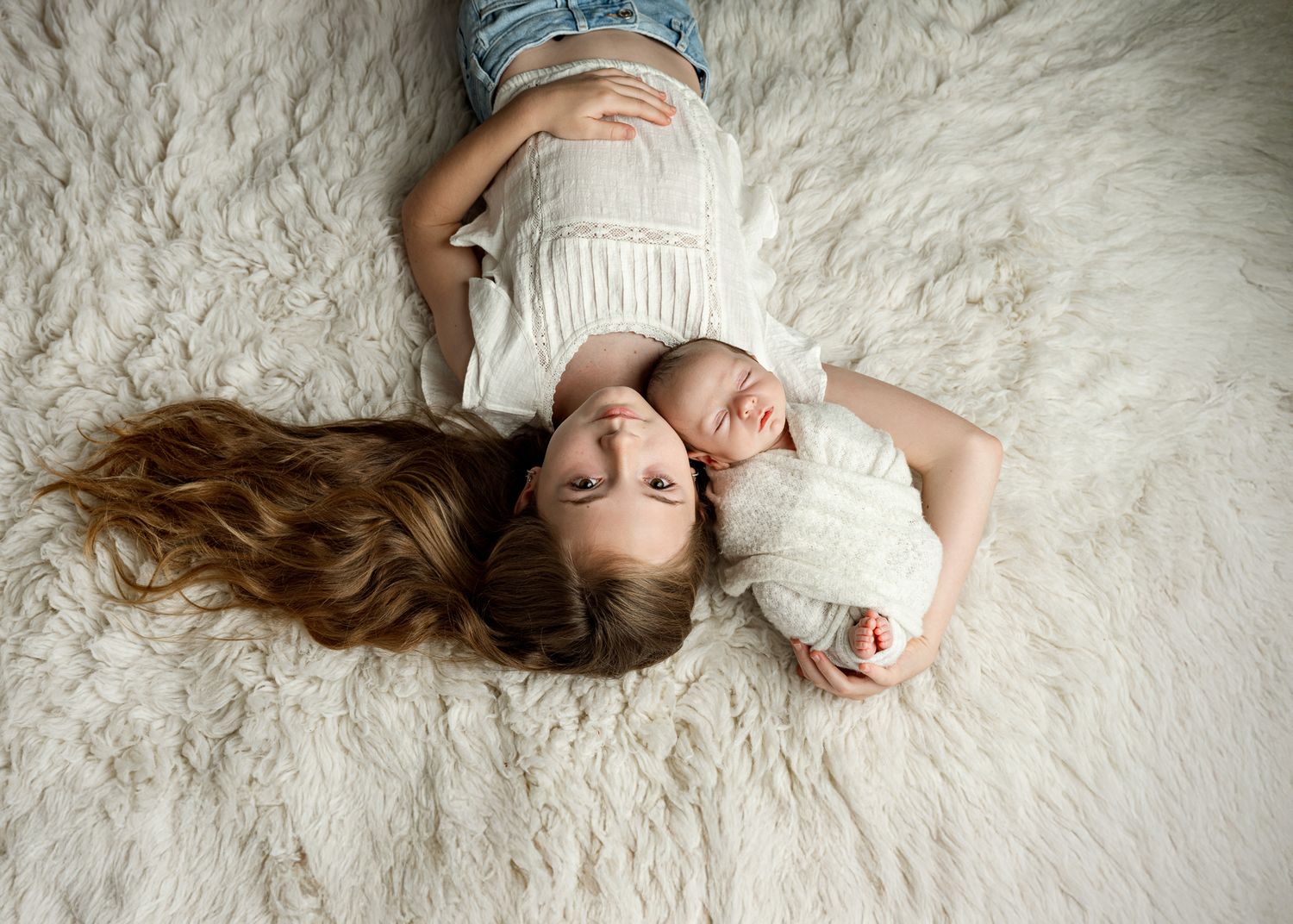 Two children lying on white furry rug looking up at camera in matching white outfits.
