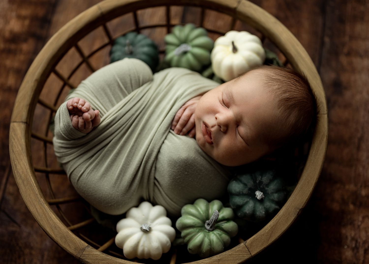 Newborn photography series showing baby sleeping in round basket with white pumpkins and sage green accents.
