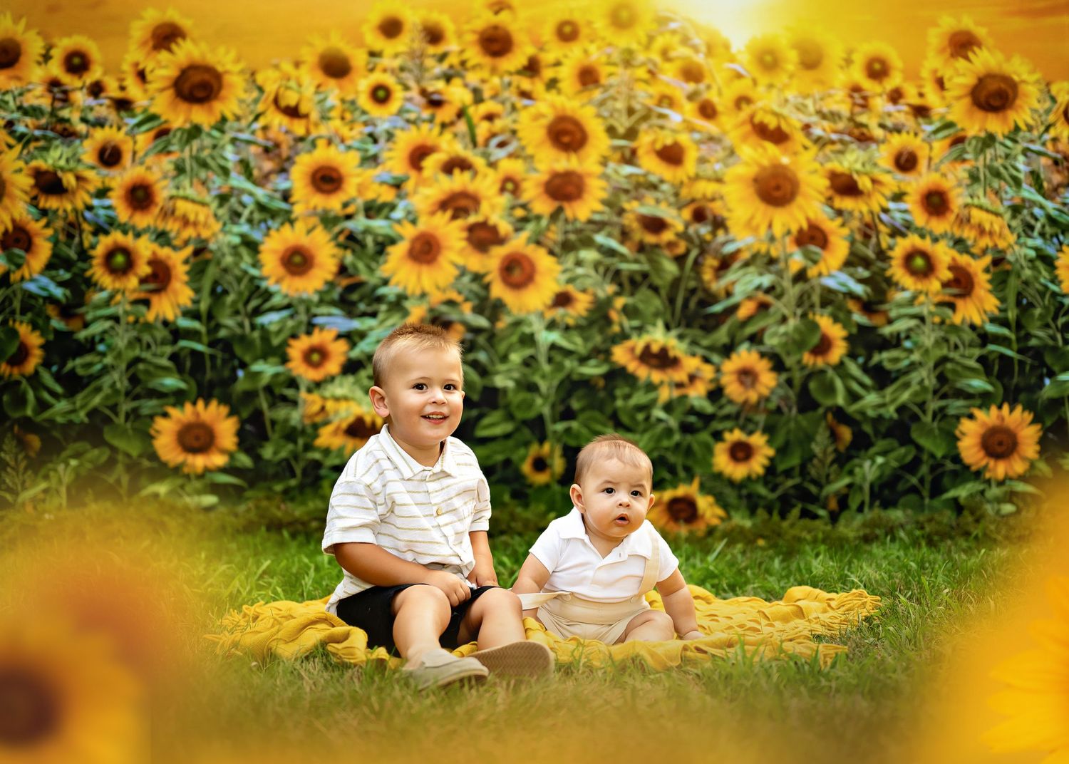 Two young children sit on a yellow blanket in front of a vibrant field of tall blooming sunflowers at sunset.