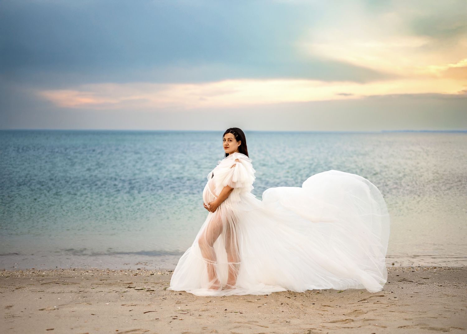 An ethereal beach maternity photoshoot at sunset with flowing white fabric and calm ocean waters in the background.