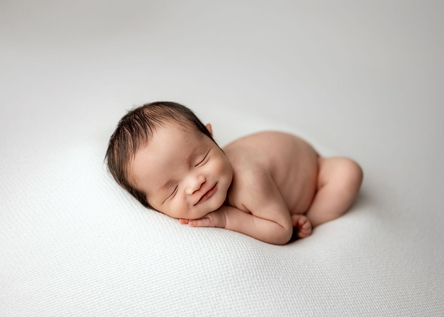A sleeping newborn baby rests peacefully on soft white fabric in a minimalist studio portrait setting.