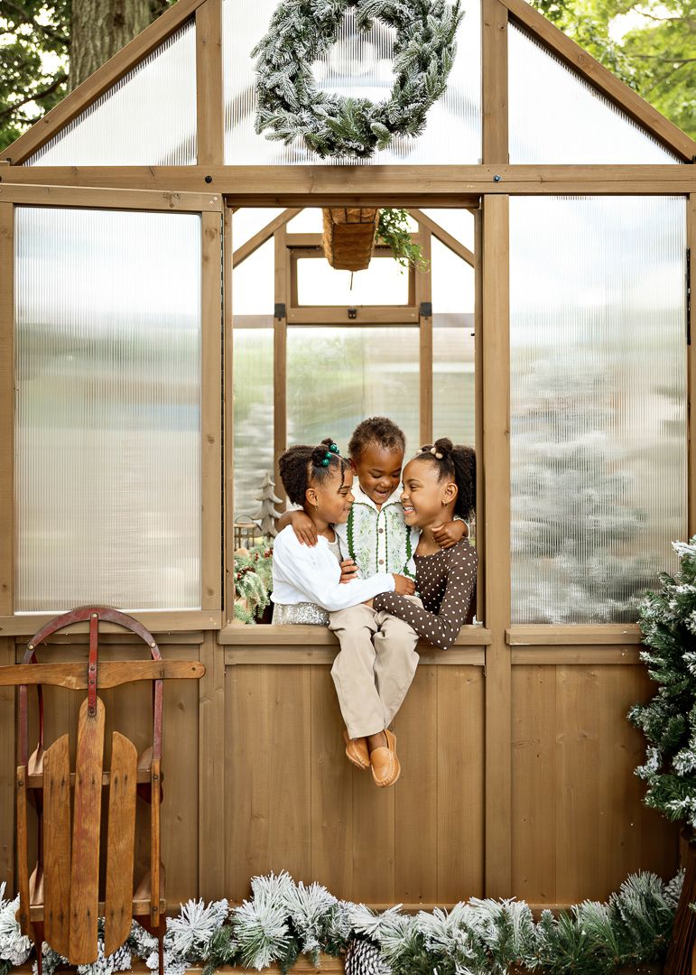 Three children sit together in a decorated greenhouse doorway with Christmas wreaths and snowy greenery.