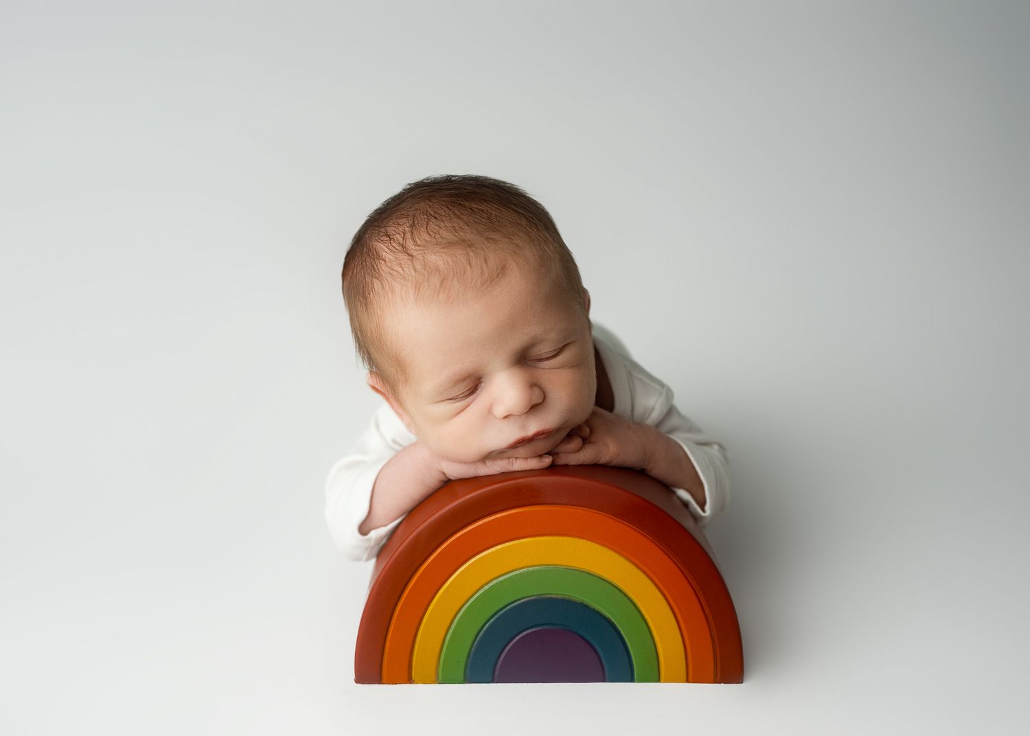 A sleeping baby rests peacefully against a colorful wooden rainbow toy on a white background.