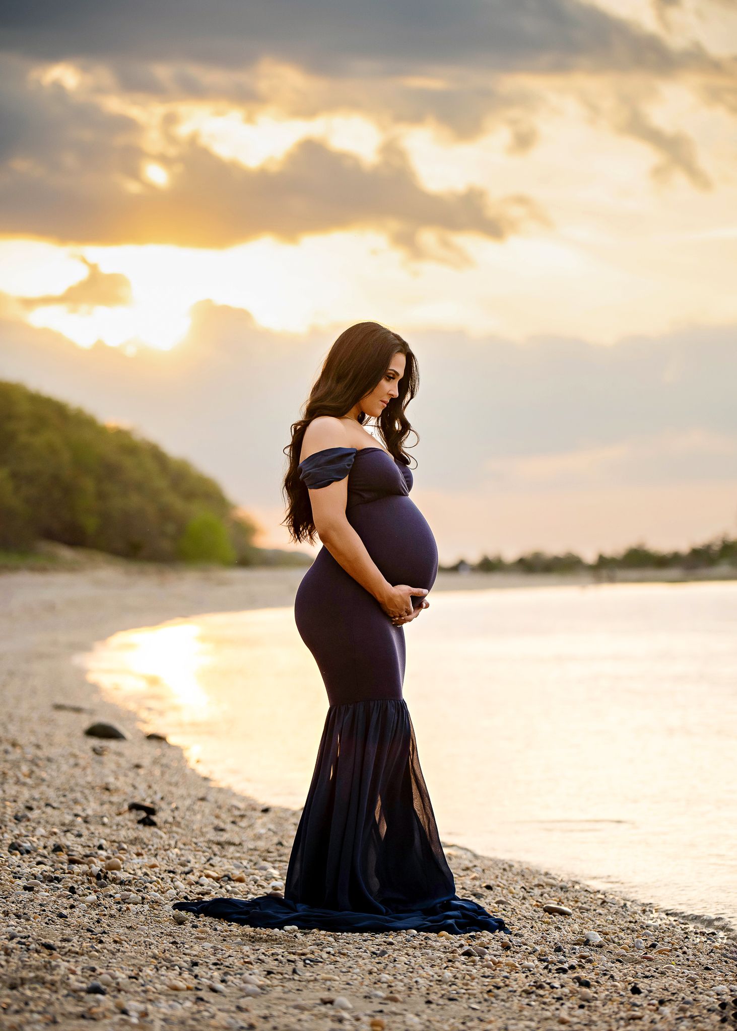 Silhouetted figure in a navy off-shoulder maternity gown poses on rocky beach at golden sunset.