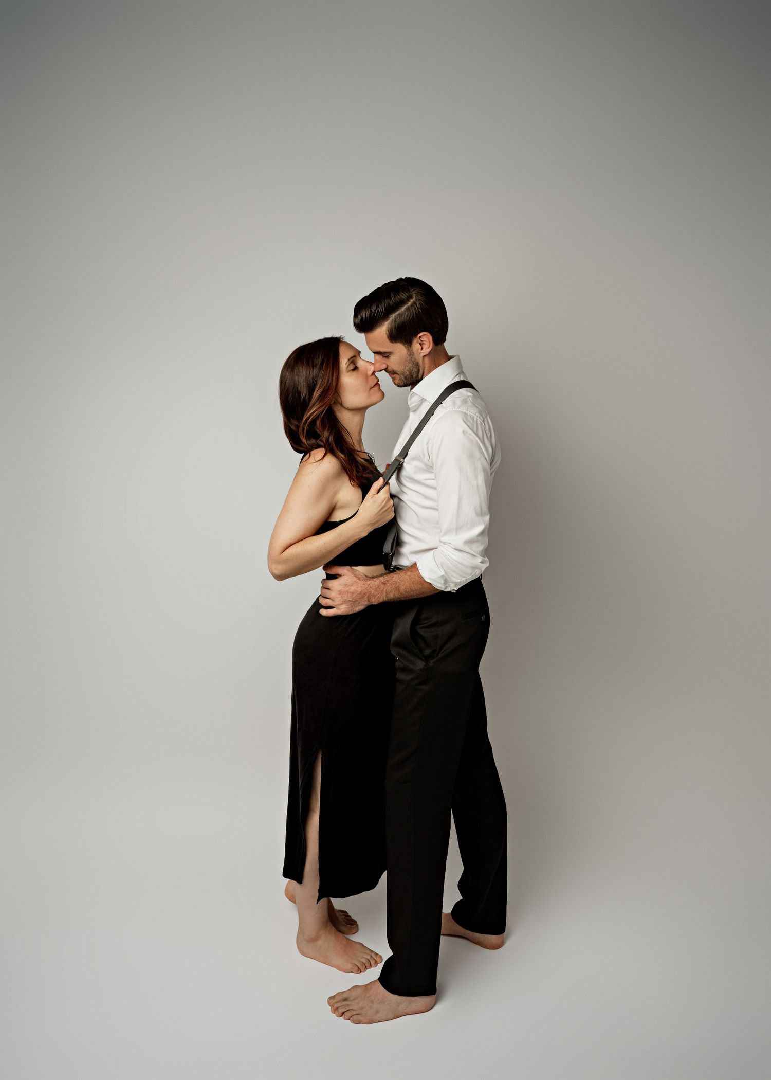 Elegantly dressed couple in black and white attire share an intimate embrace against neutral backdrop.