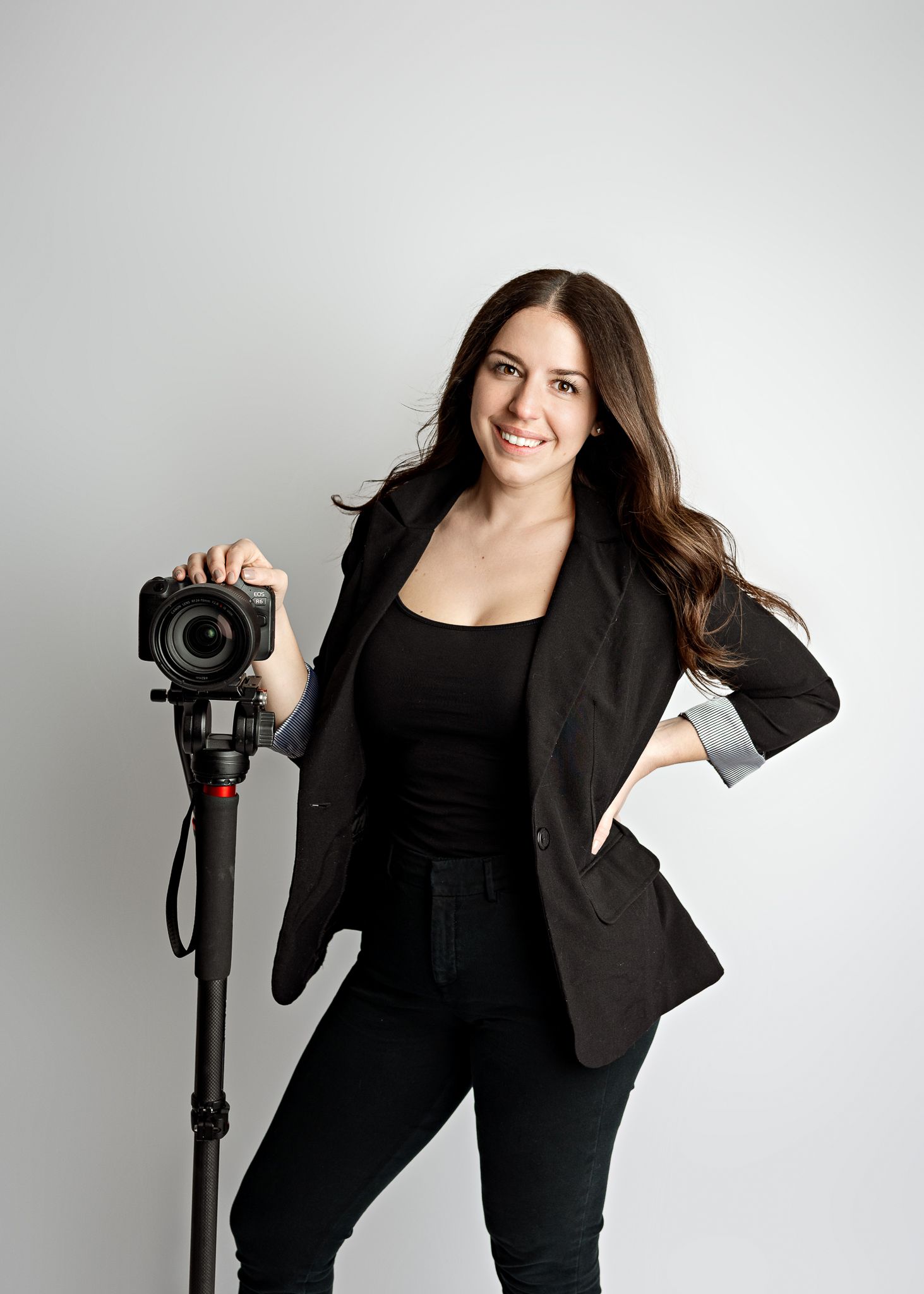 Professional photographer wearing all black business attire poses with camera on tripod against light gray background.
