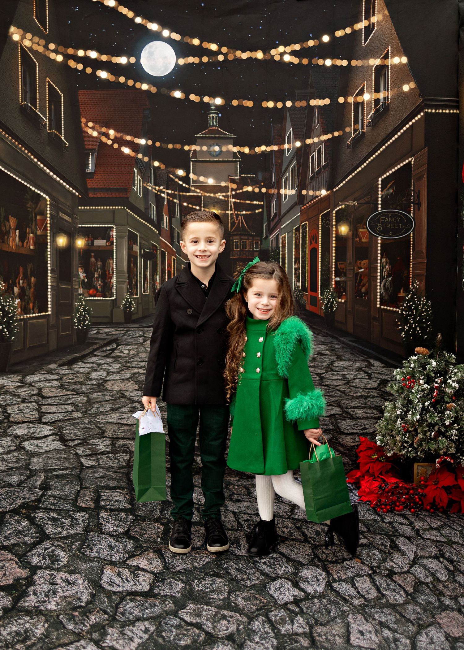A cobblestone street decorated with string lights and holiday decor features two young children in formal winter attire at night.