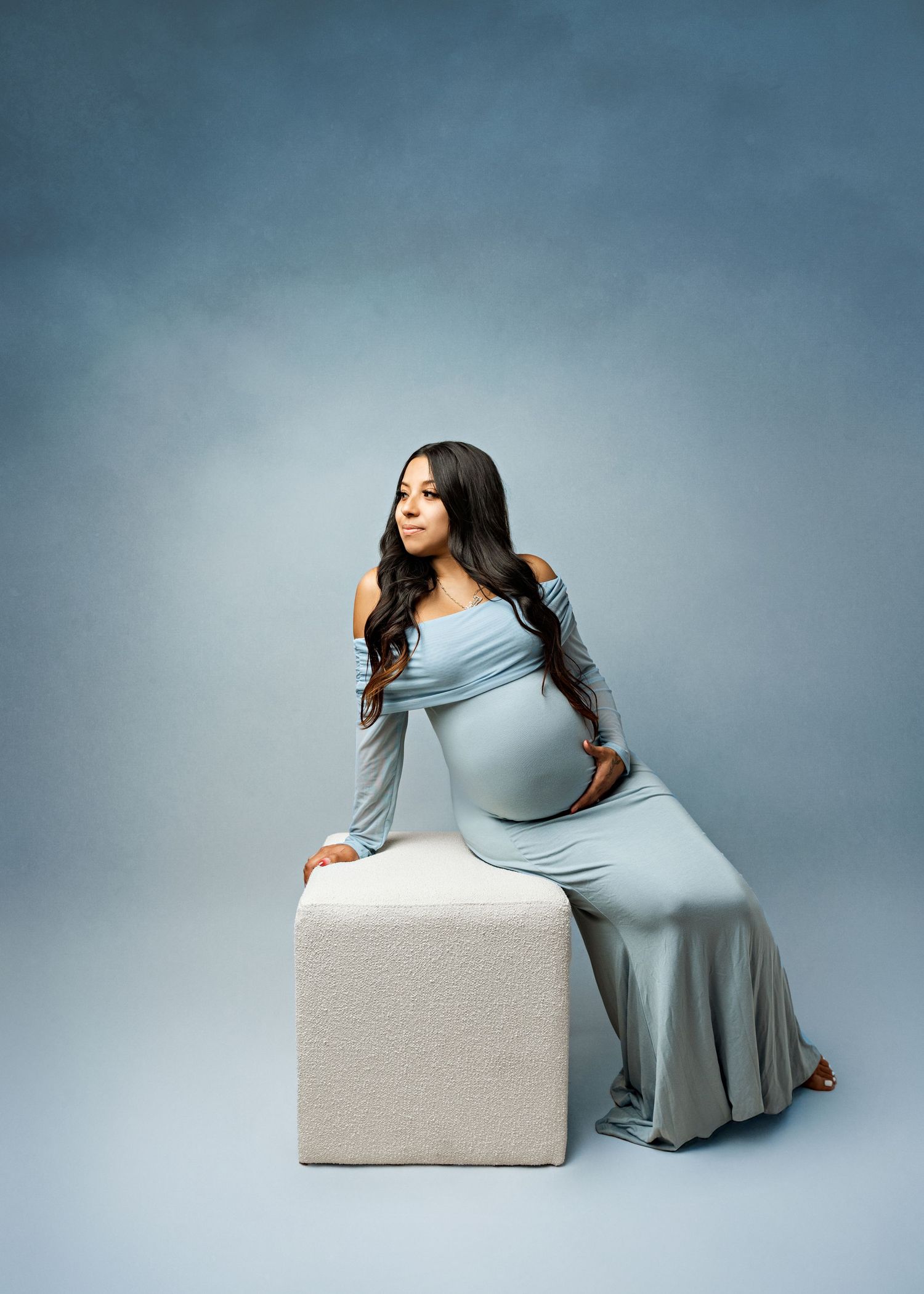 A figure in a flowing light blue maternity dress poses elegantly against a white cube against a gray studio backdrop.