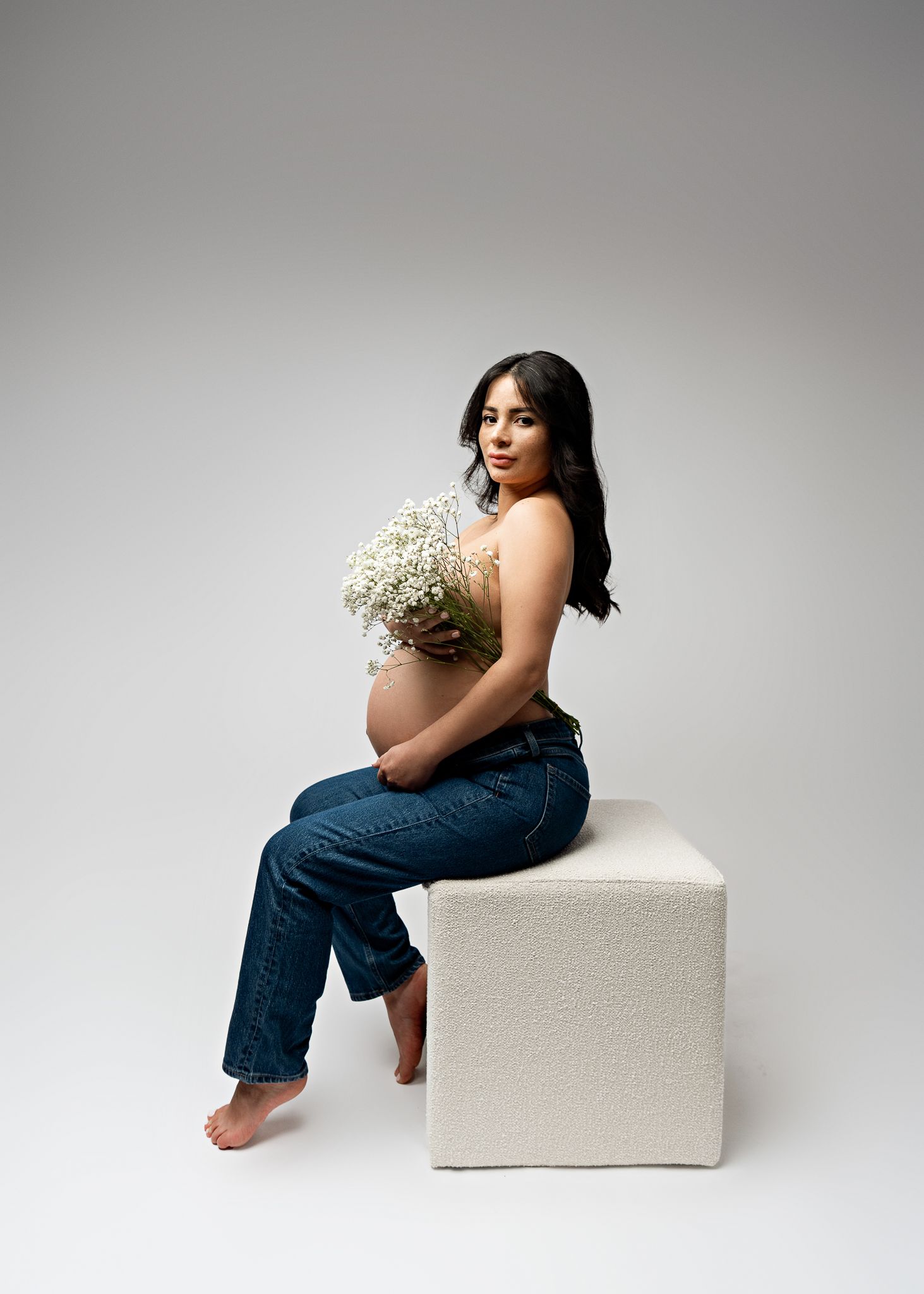 Expectant mother in blue jeans sits on white cube holding baby's breath flowers in studio setting.