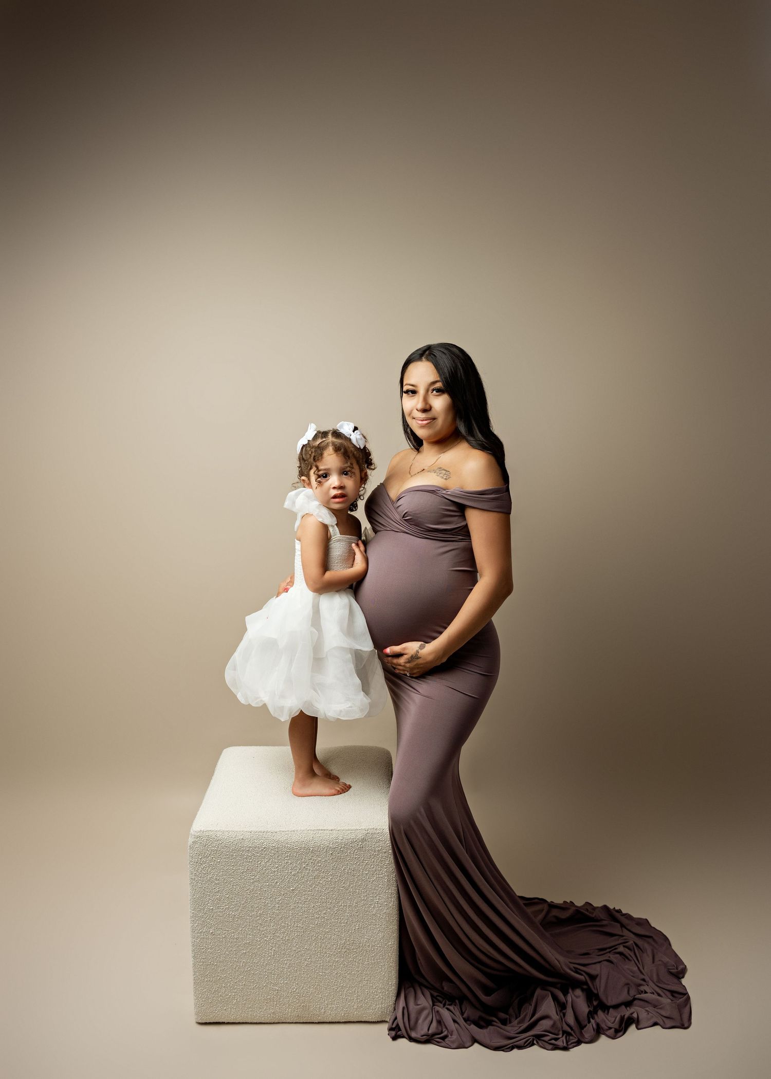 Elegant maternity photo of two figures in coordinating dresses posing together against a neutral studio backdrop.