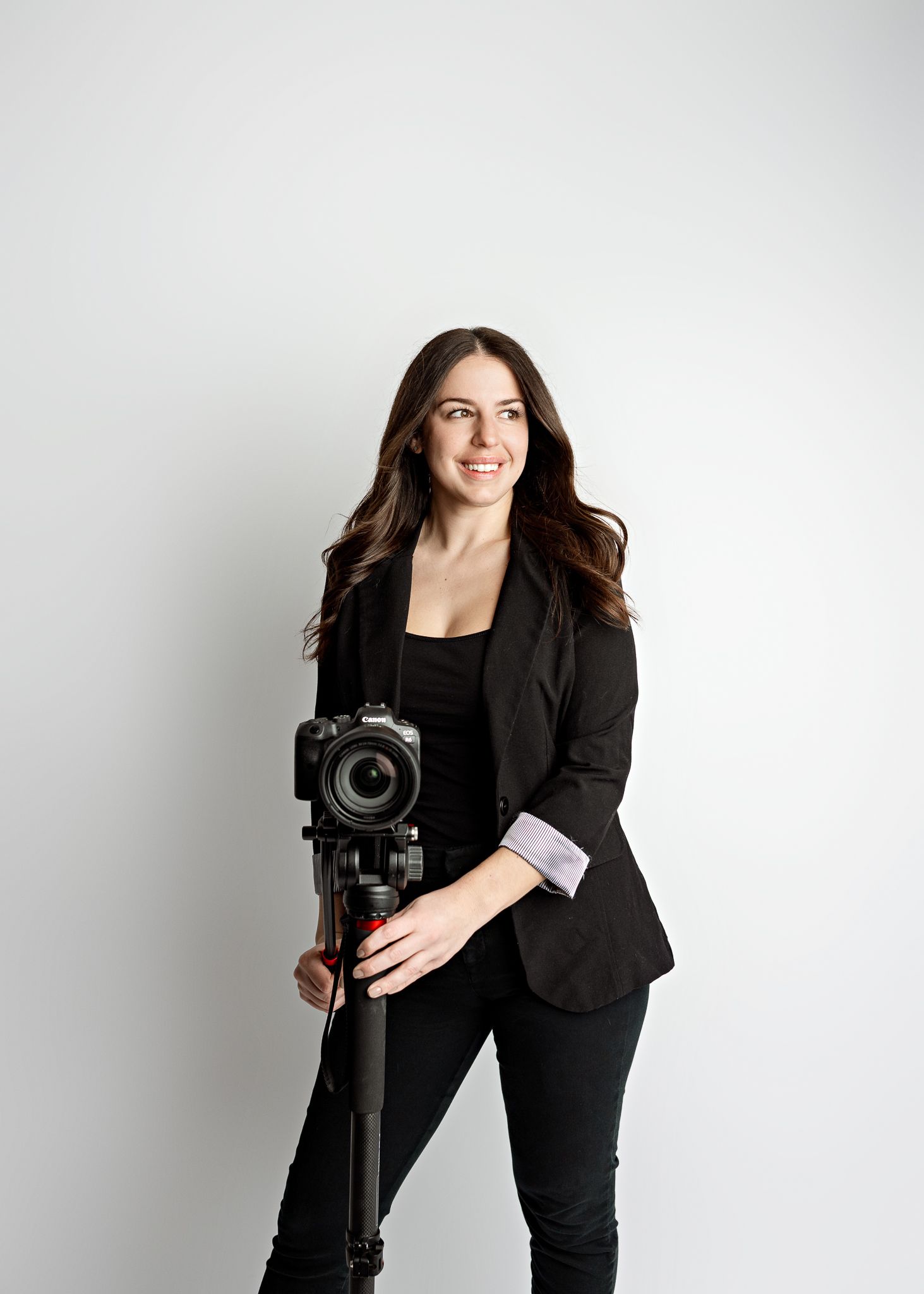 Professional photographer in black outfit poses with camera equipment against white studio backdrop.