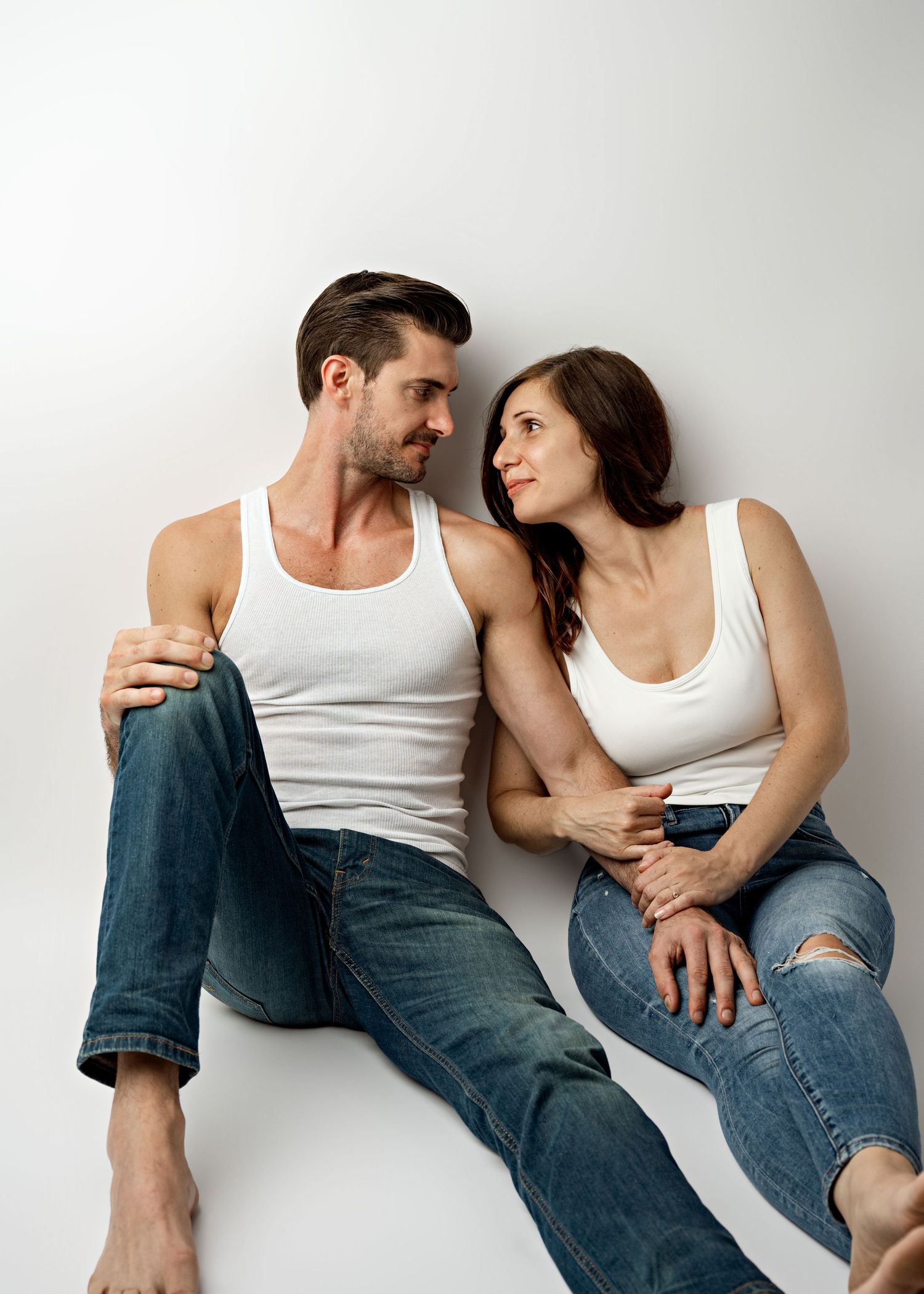 A couple wearing white tank tops and blue jeans sit casually together against a light background.