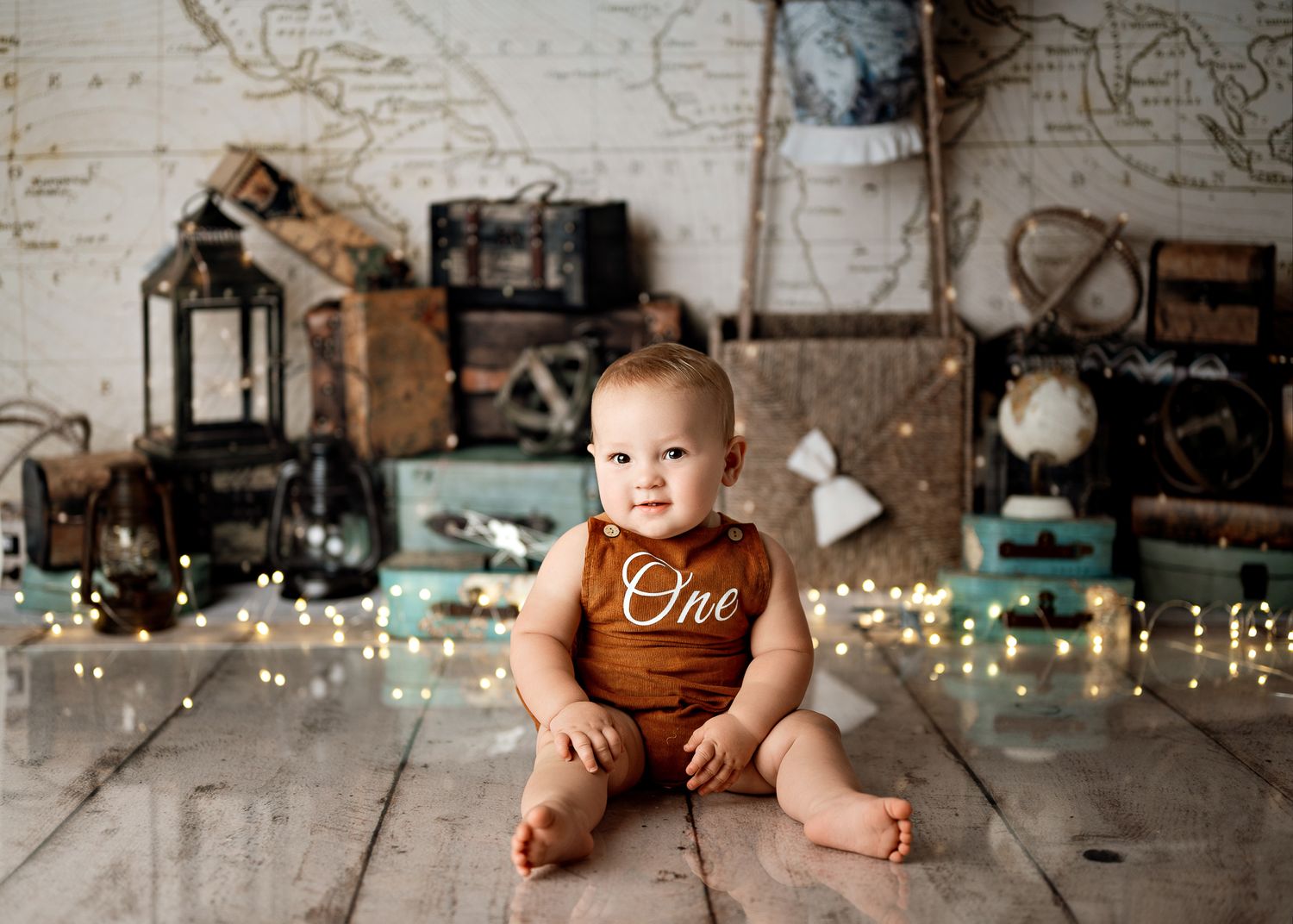 Small baby wearing a brown romper sits on wooden floor with vintage decor and twinkling string lights in background.