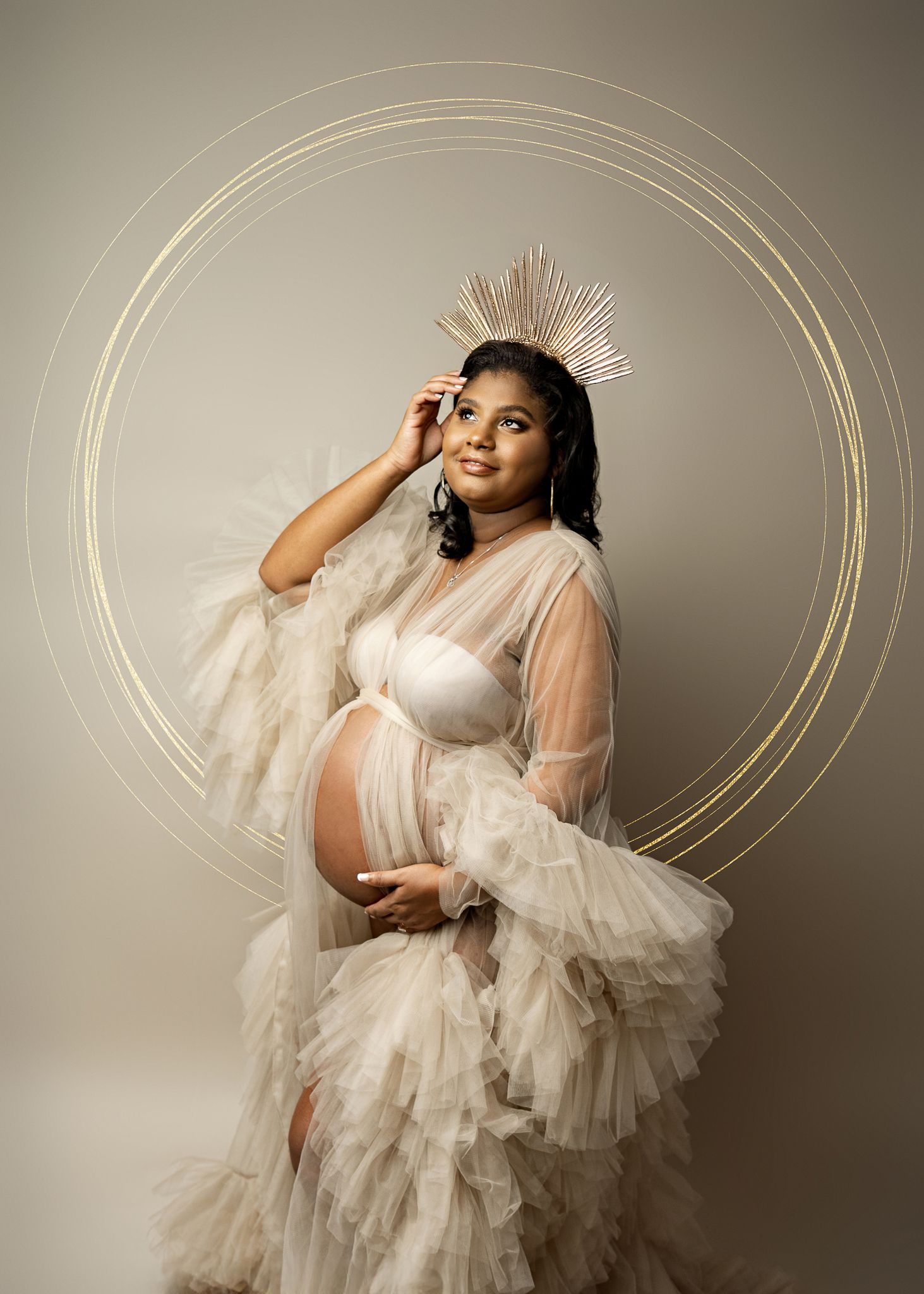 An ethereal maternity portrait featuring a sheer tulle gown and radiant crown headpiece against a neutral backdrop.