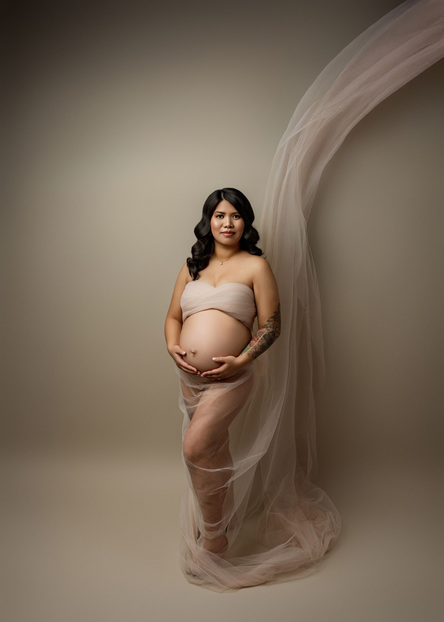 A series of ethereal maternity photos showing a pregnant figure draped in flowing beige tulle fabric against a neutral backdrop.