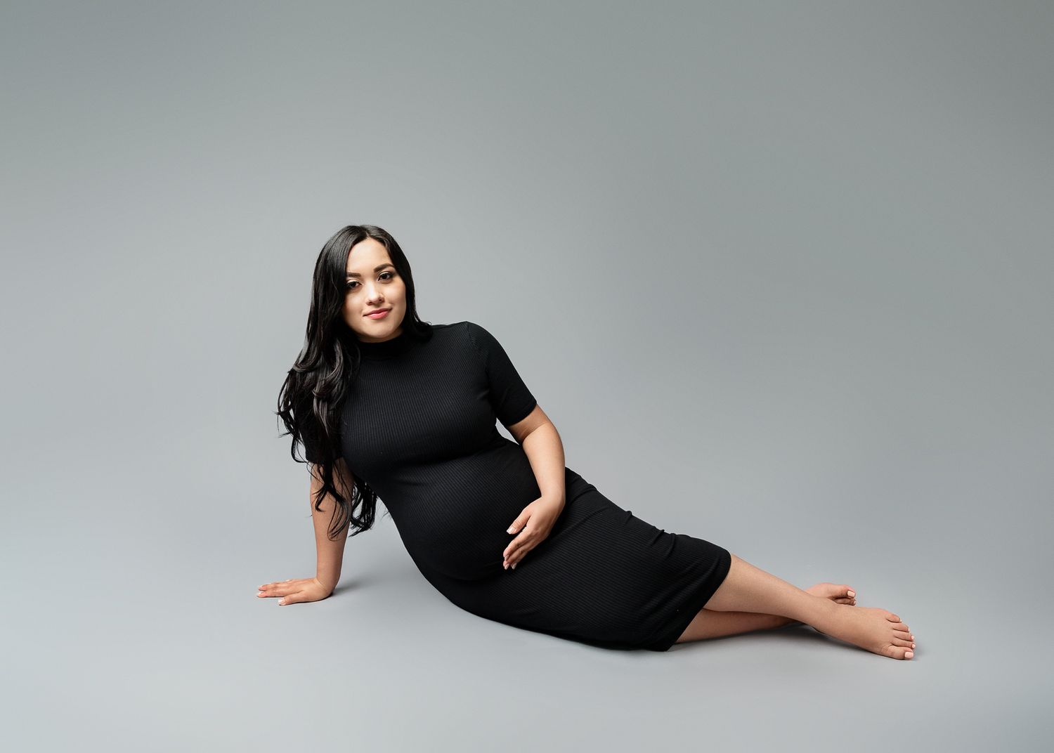 A person in a black short-sleeved dress reclines on a gray studio floor against a neutral background.