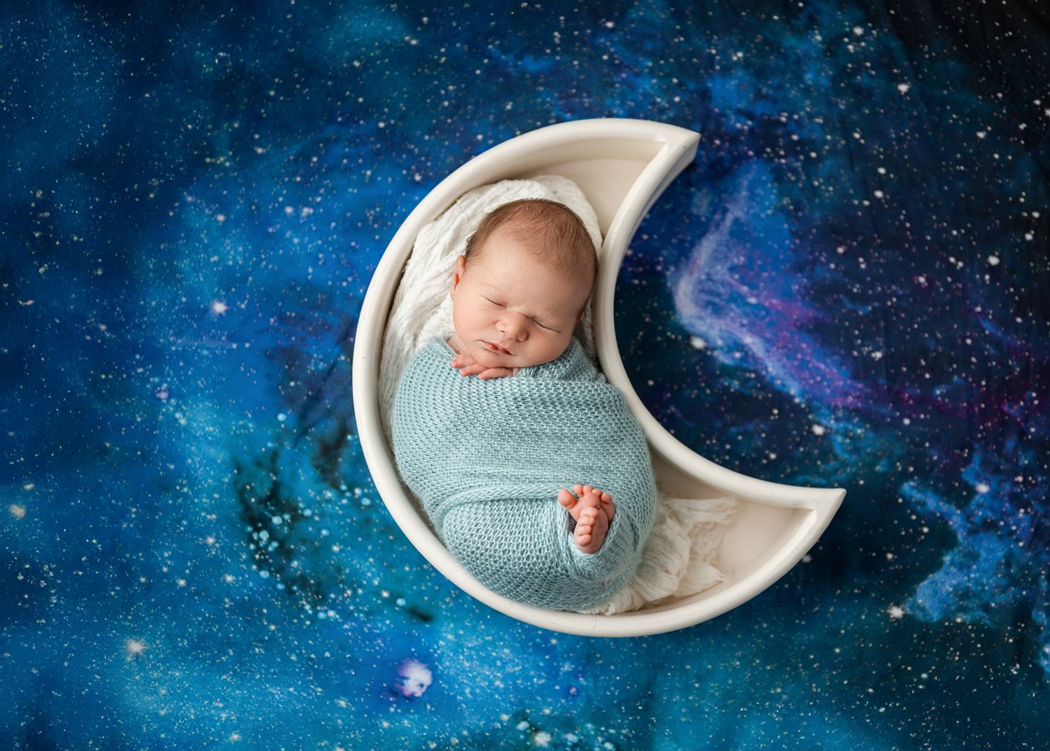 A baby sleeps peacefully in a crescent moon shaped prop against a starry blue galaxy background.