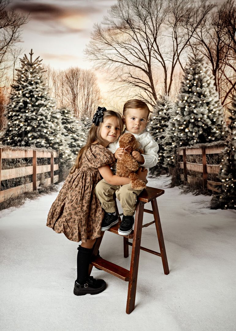 Two young siblings in holiday attire posing together on stool with teddy bear in snowy Christmas tree setting.