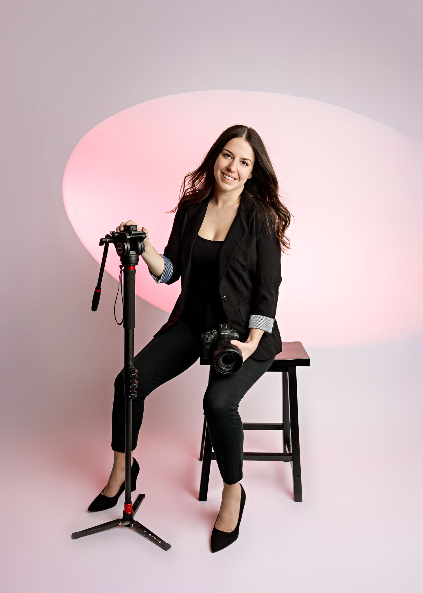 Professional photographer in all black attire sits on stool with camera and tripod against pink backdrop.