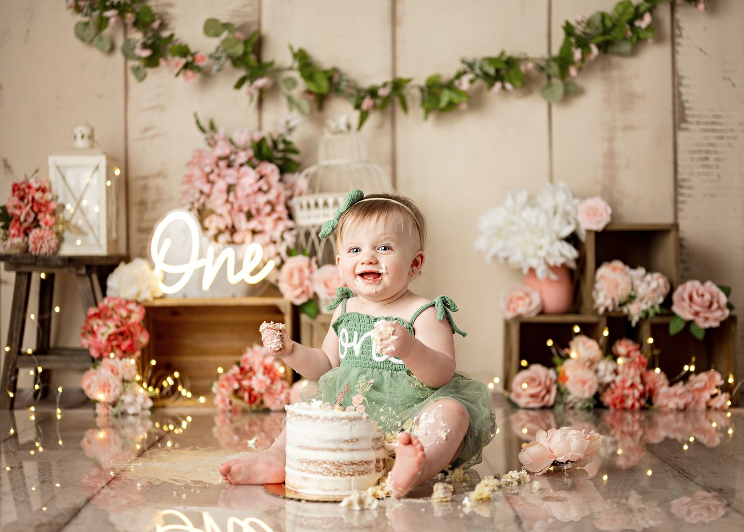 Cake smash photography setup with floral decorations, greenery garland, and vintage decor in soft pink and cream tones.