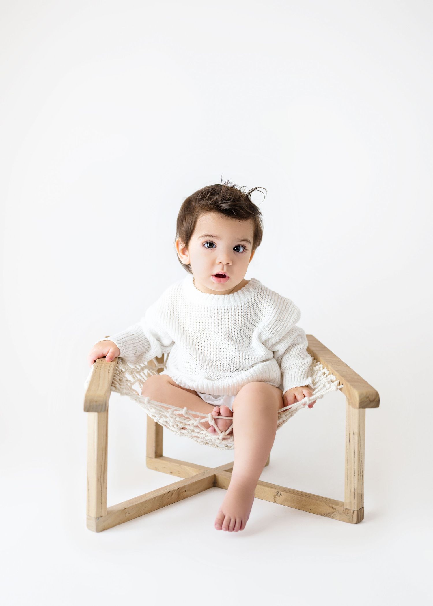 Small child wearing a white sweater sits in a modern wooden chair with woven seat against a light background.