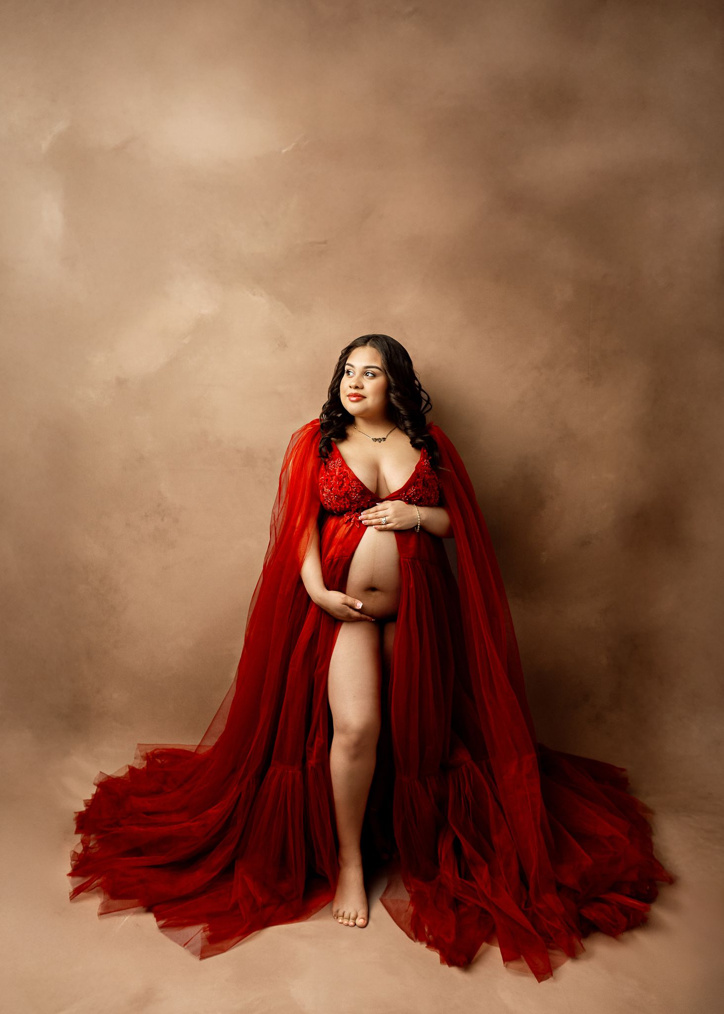 A person wearing a flowing red cape poses gracefully against a sepia-toned backdrop in an artistic maternity photoshoot.