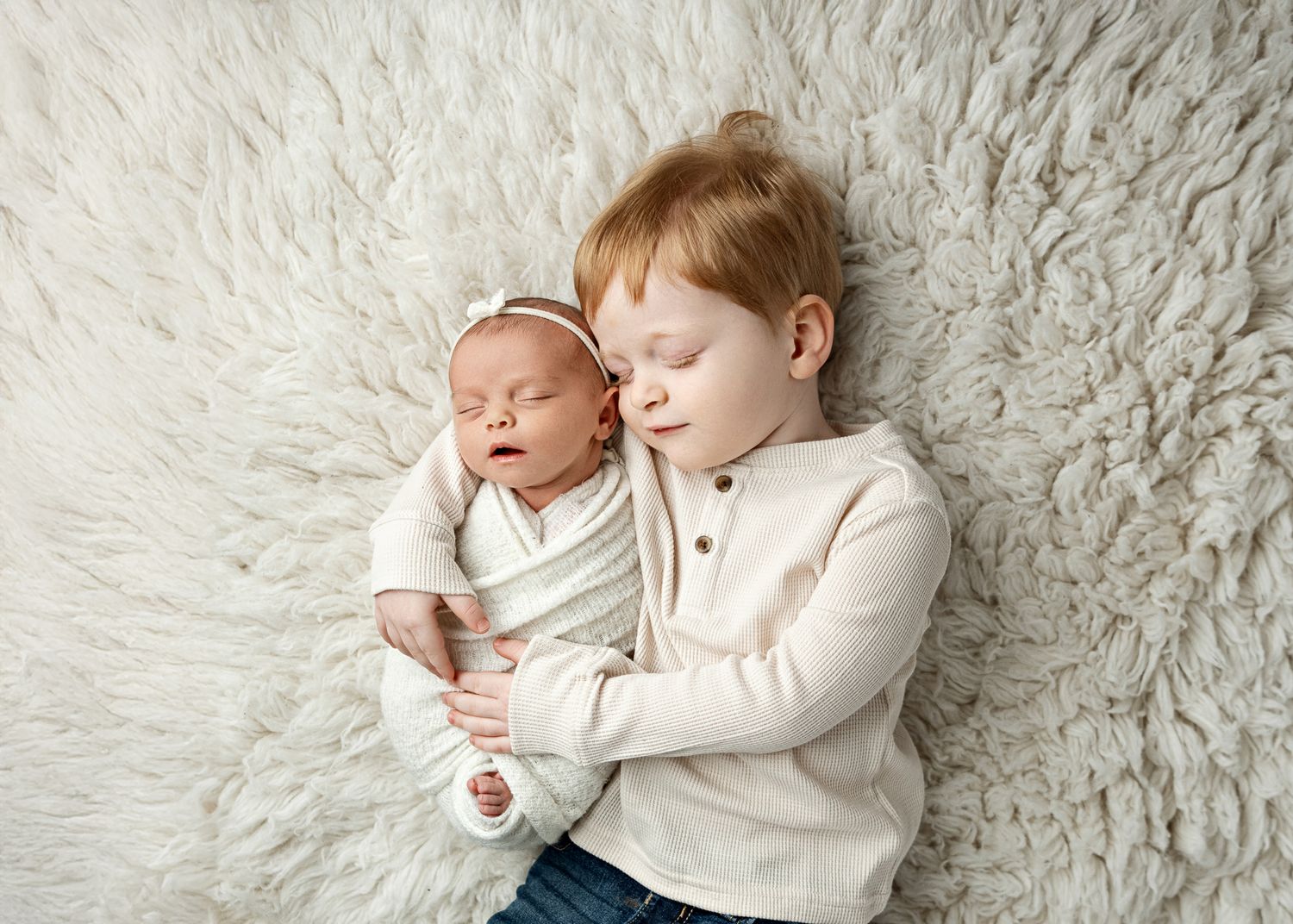 Two children resting peacefully on soft white textured bedding wearing matching cream colored outfits.