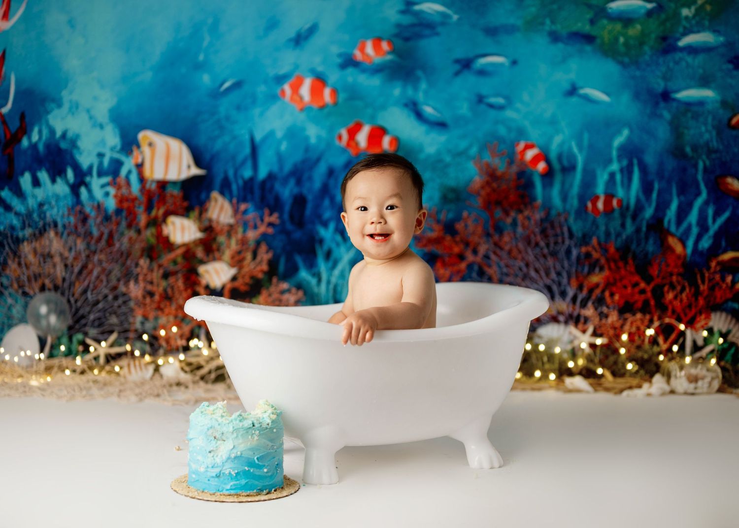 Baby bathtub photo session with underwater ocean backdrop and clownfish decorations plus blue smash cake.