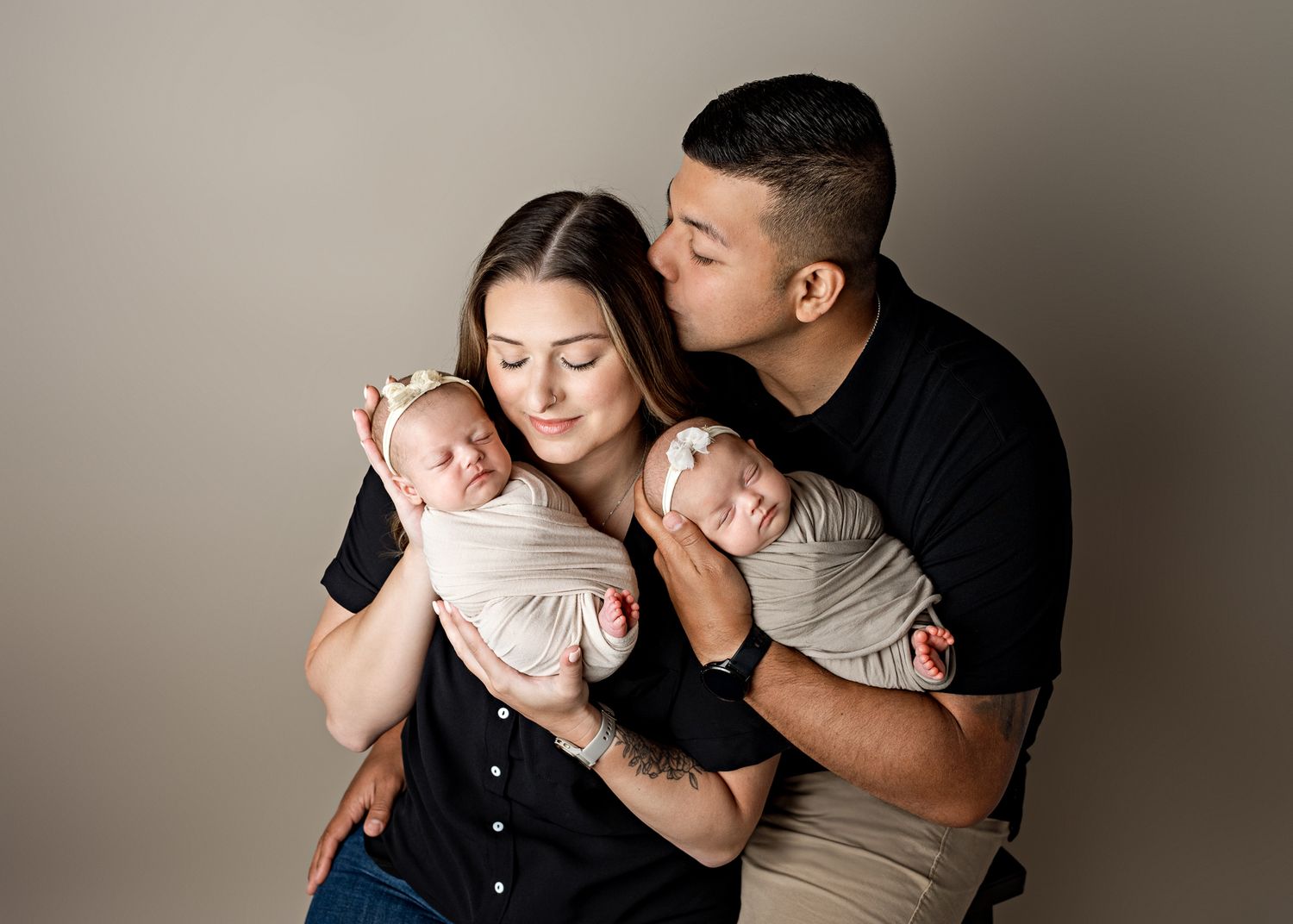 Couple in black and beige clothing holding their newborn baby in a tender embrace against neutral backdrop.
