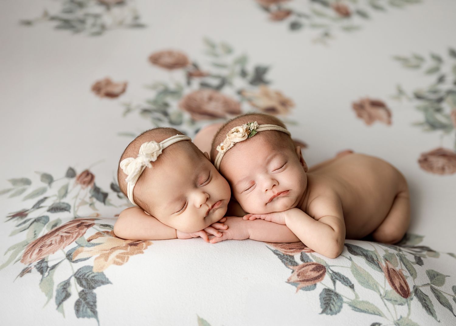 Two sleeping newborns wearing delicate headbands rest together on a floral patterned blanket.