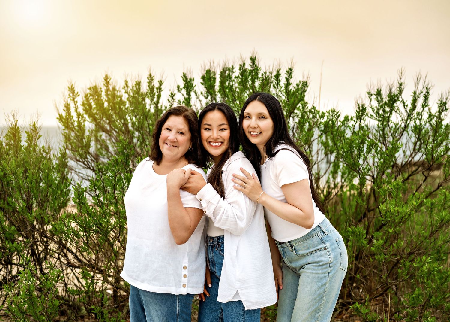 Three people in casual outfits laughing and embracing while standing in front of green bushes outdoors.
