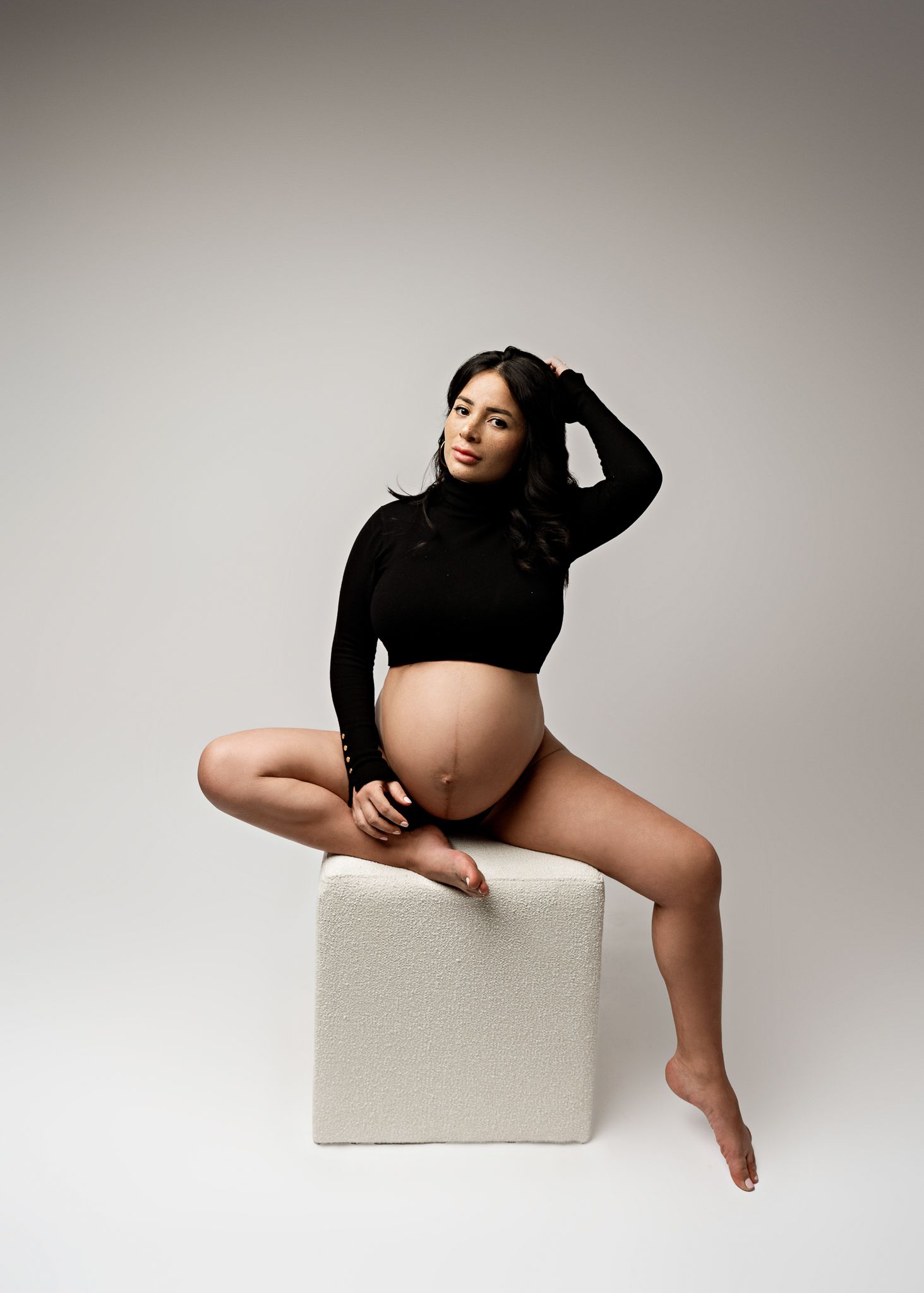 Artistic maternity portrait of woman in black crop top posing on white cube against neutral backdrop on Long Island, NY