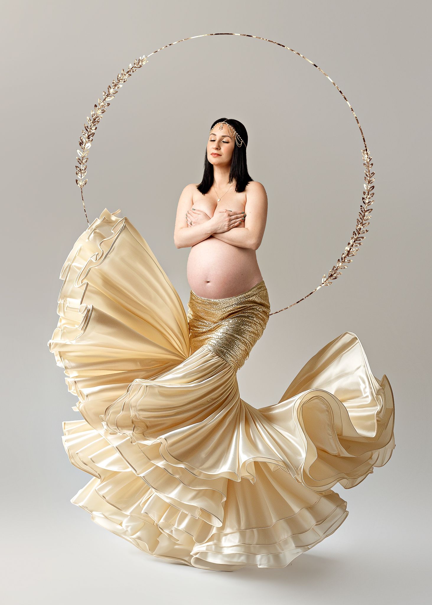 Ethereal maternity photo featuring a flowing cream-colored dress and circular floral hoop against a gray backdrop.