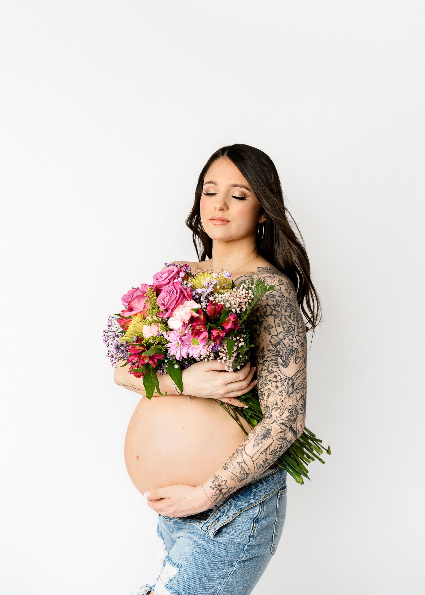 A pregnant woman with tattoo sleeves holds a vibrant bouquet of pink flowers against a white background.